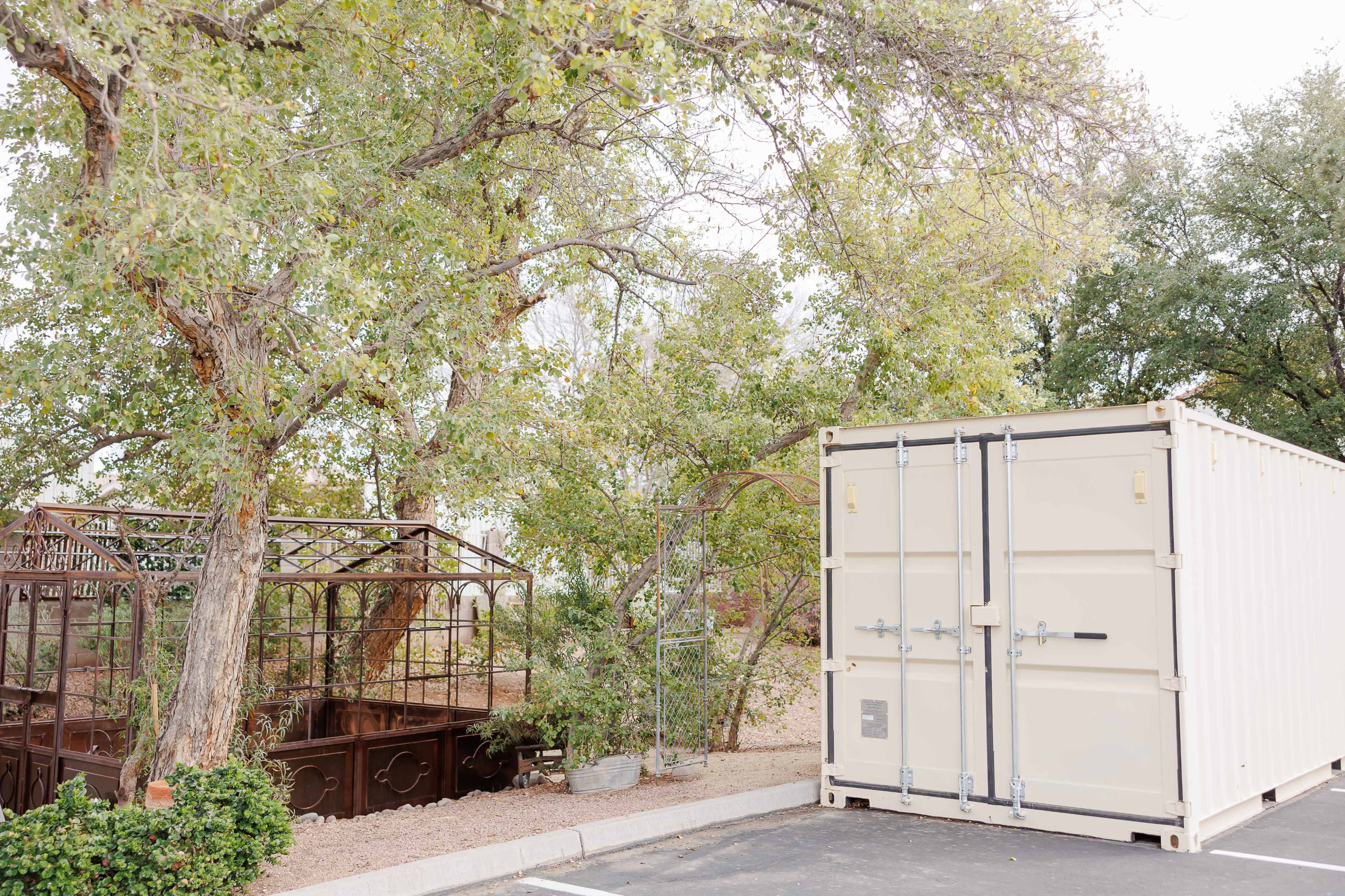 A large shipping container is displayed next to a metal greenhouse surrounded by trees in a parking area.