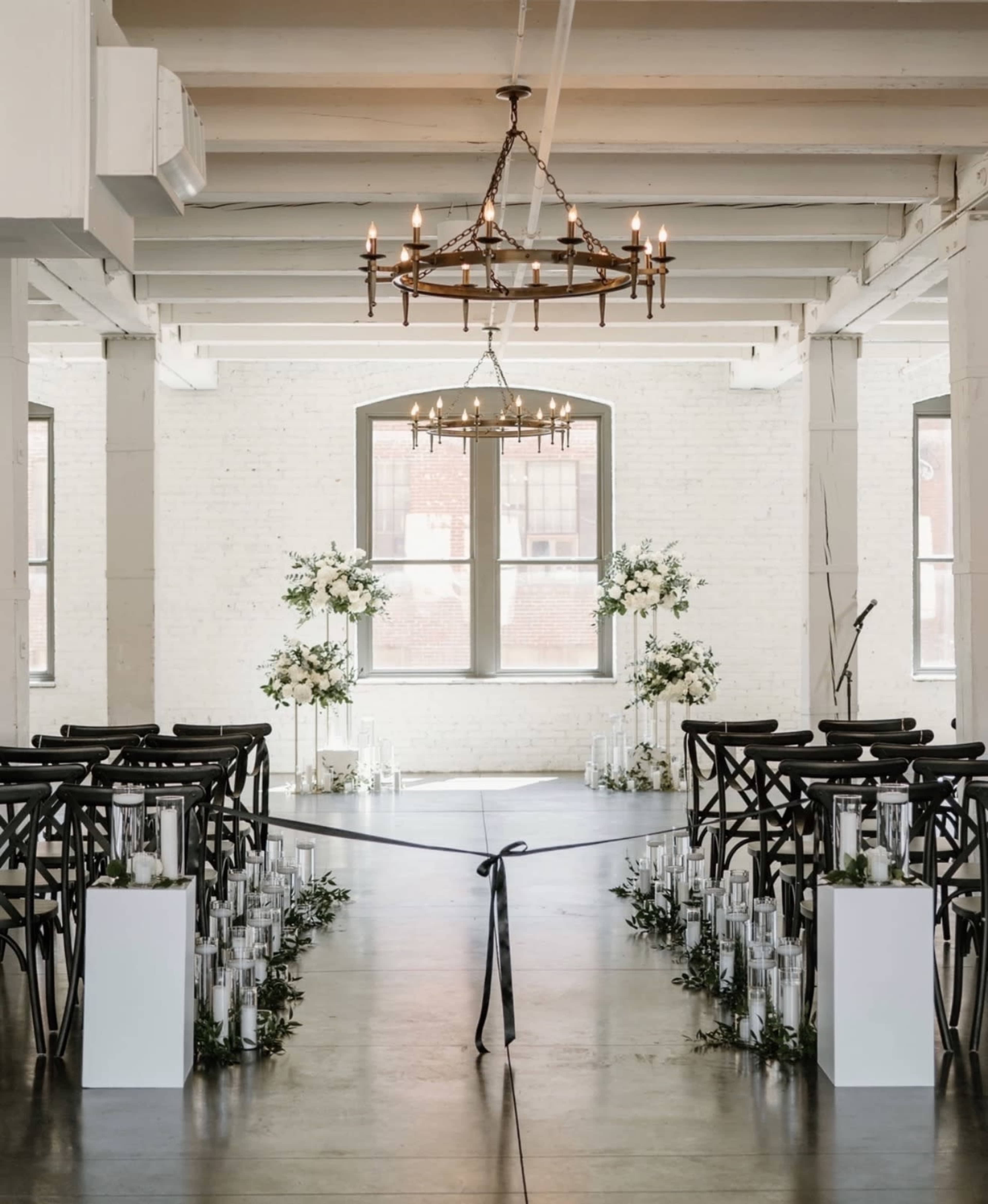 An indoor wedding ceremony setup features rows of black wooden chairs flanking a central aisle lined with white pillars and glass vases, all beneath a chandelier and surrounded by floral arrangements.