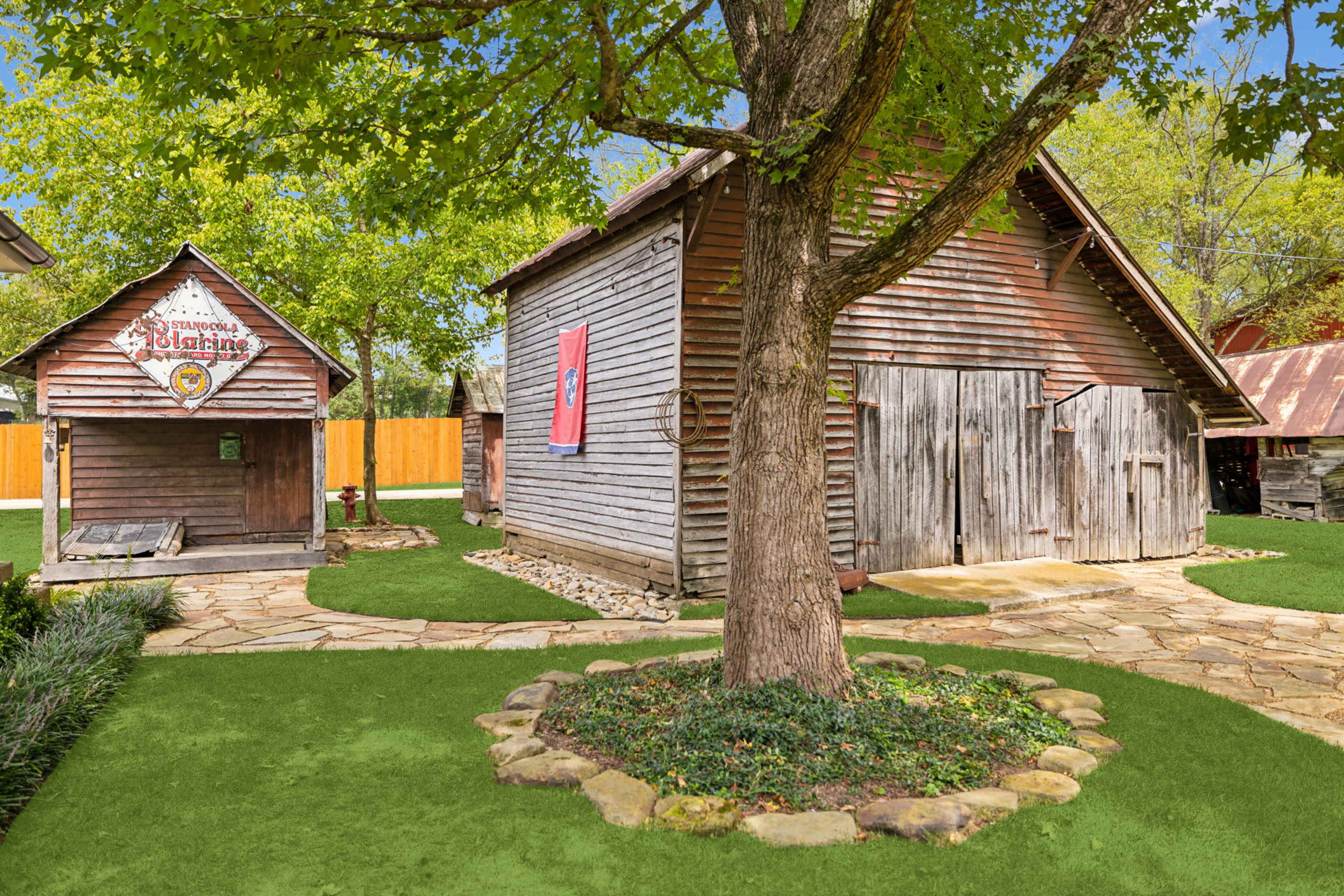 The image depicts a rustic outdoor scene featuring two wooden structures, a tree, and a stone pathway surrounded by artificial grass.