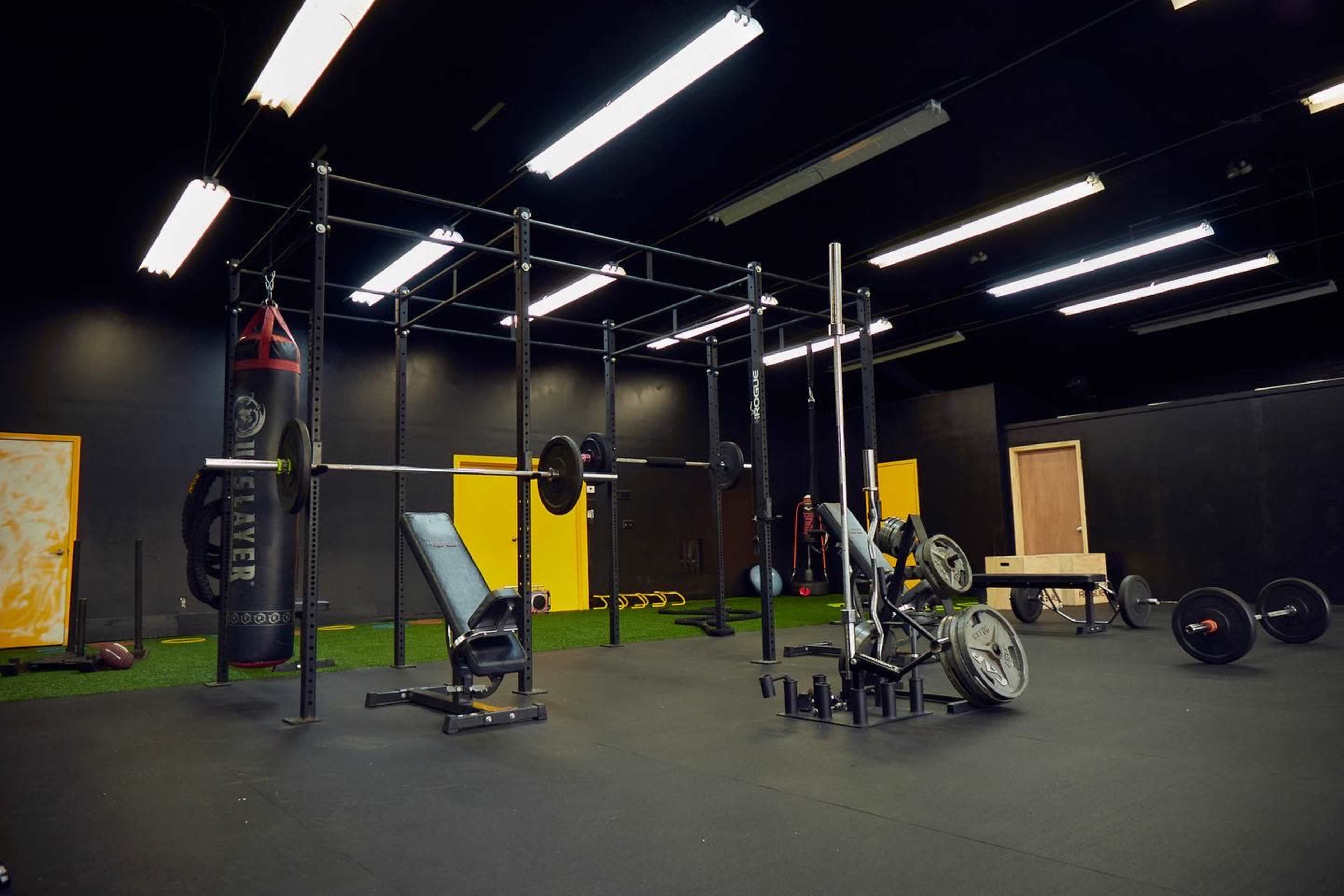 A gym interior featuring a weightlifting area with a barbell, a bench, and various pieces of fitness equipment against a black backdrop.