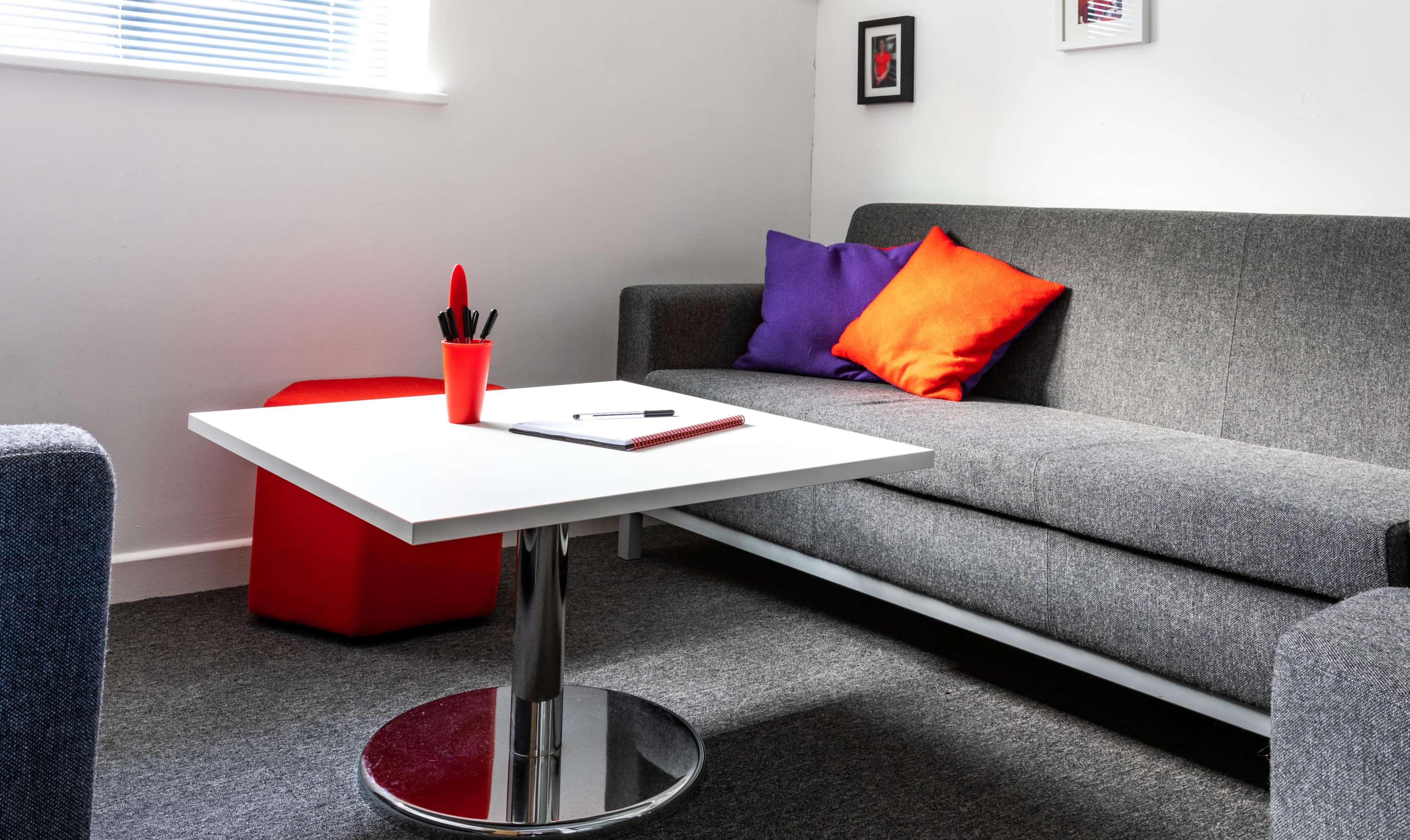 The image features a modern living space with a gray sofa, colorful cushions, a round coffee table with a notebook and pen, and a red cube-shaped stool.
