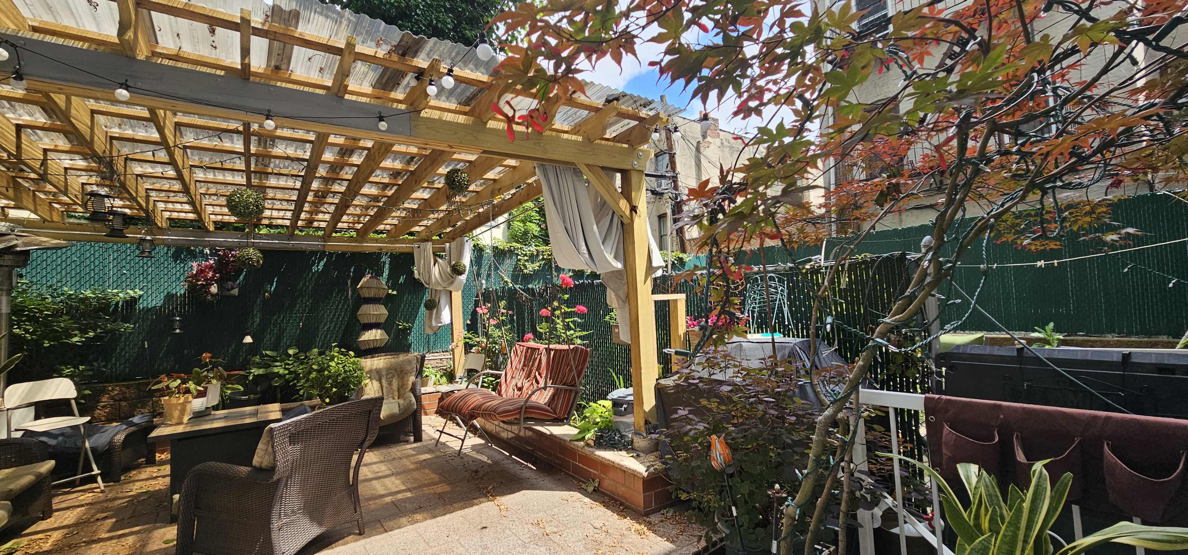 A backyard patio is sheltered by a wooden pergola adorned with hanging plants and string lights, featuring seating areas and surrounding greenery.