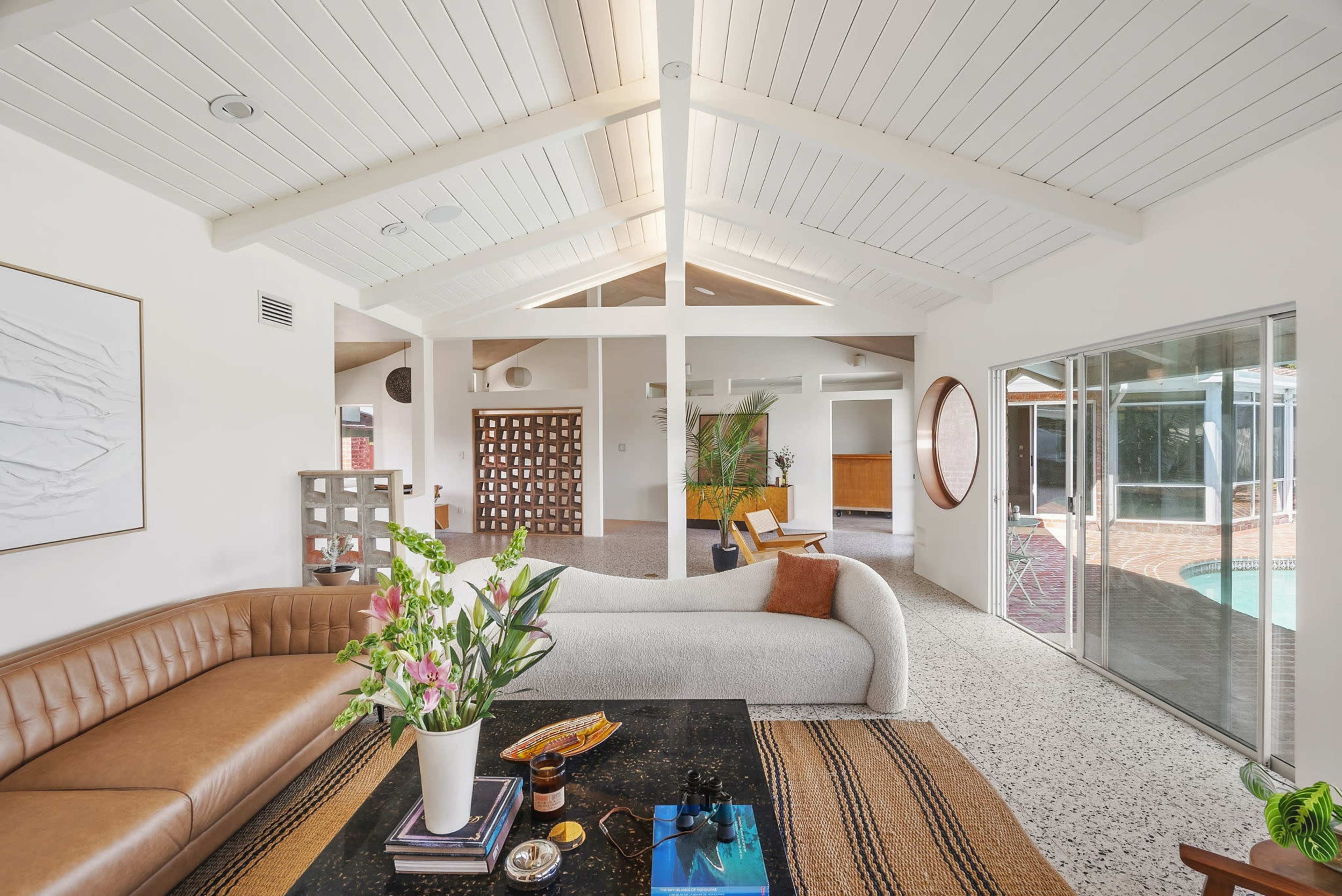 The image shows a modern, airy living space with a beige sofa, a coffee table, and a view of a pool through large glass doors.