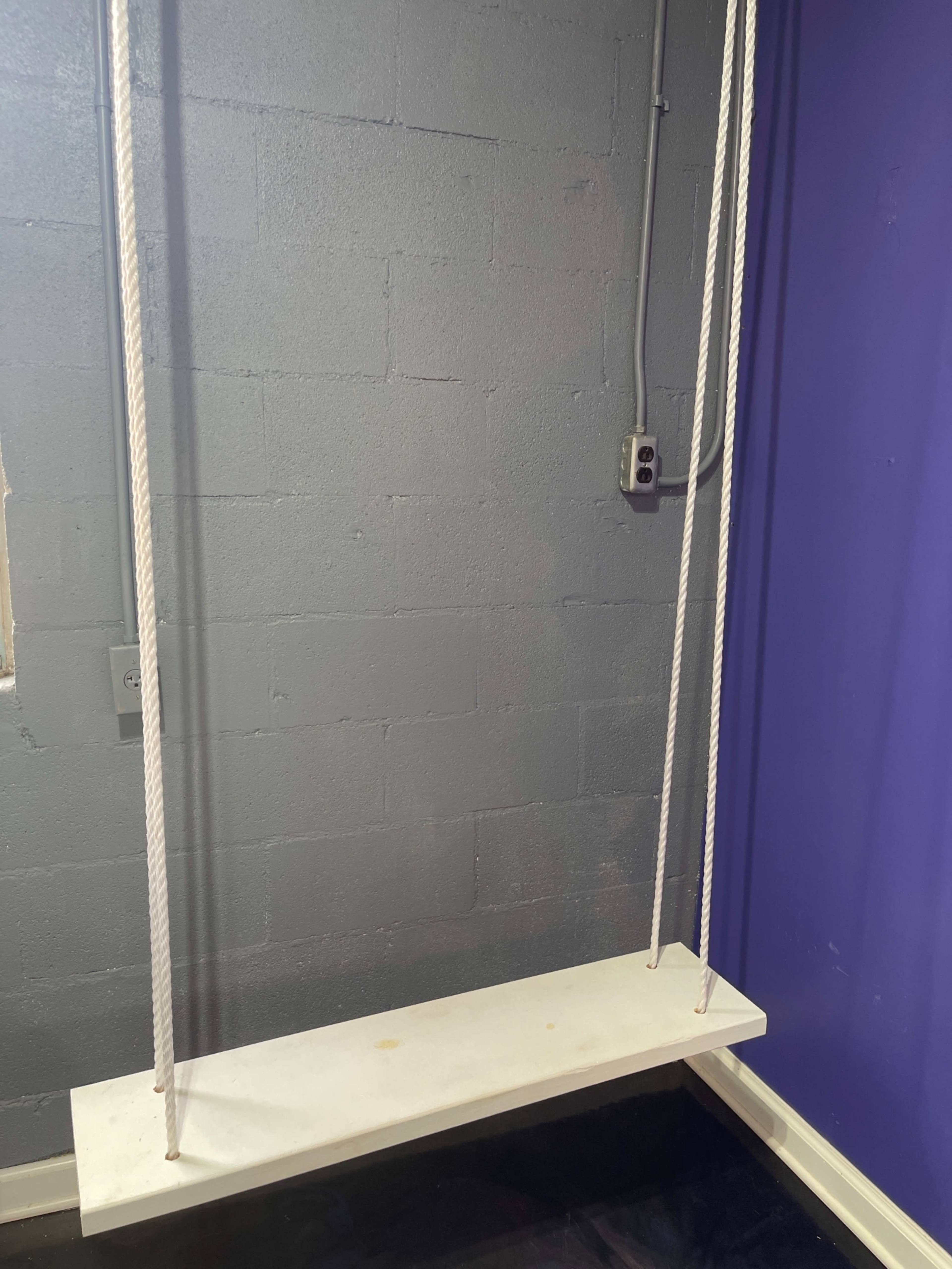 A wooden swing suspended by ropes hangs against a gray wall in a room with purple accents.