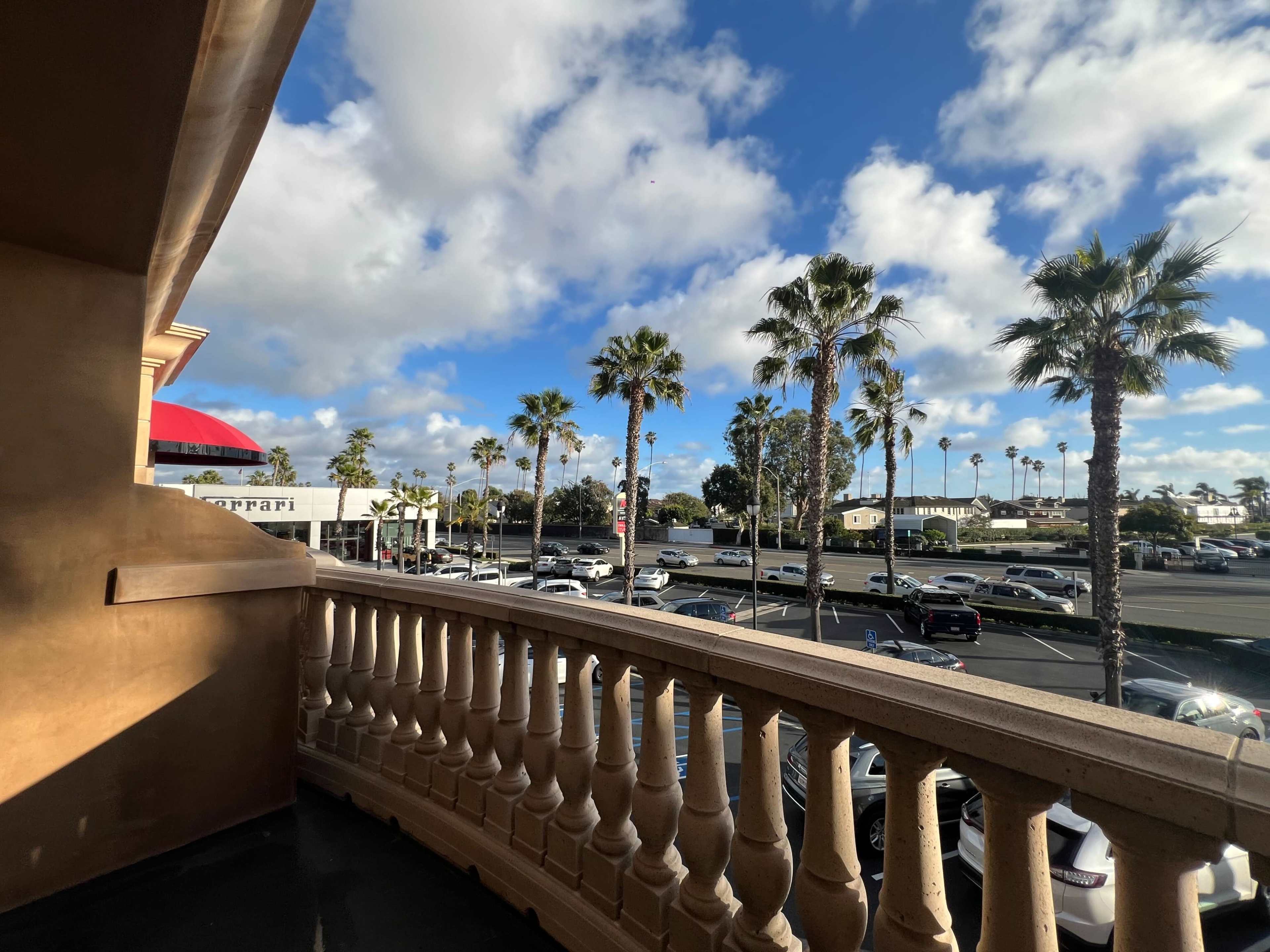 A view from a balcony shows palm trees and parked cars in a lot under a partly cloudy sky.