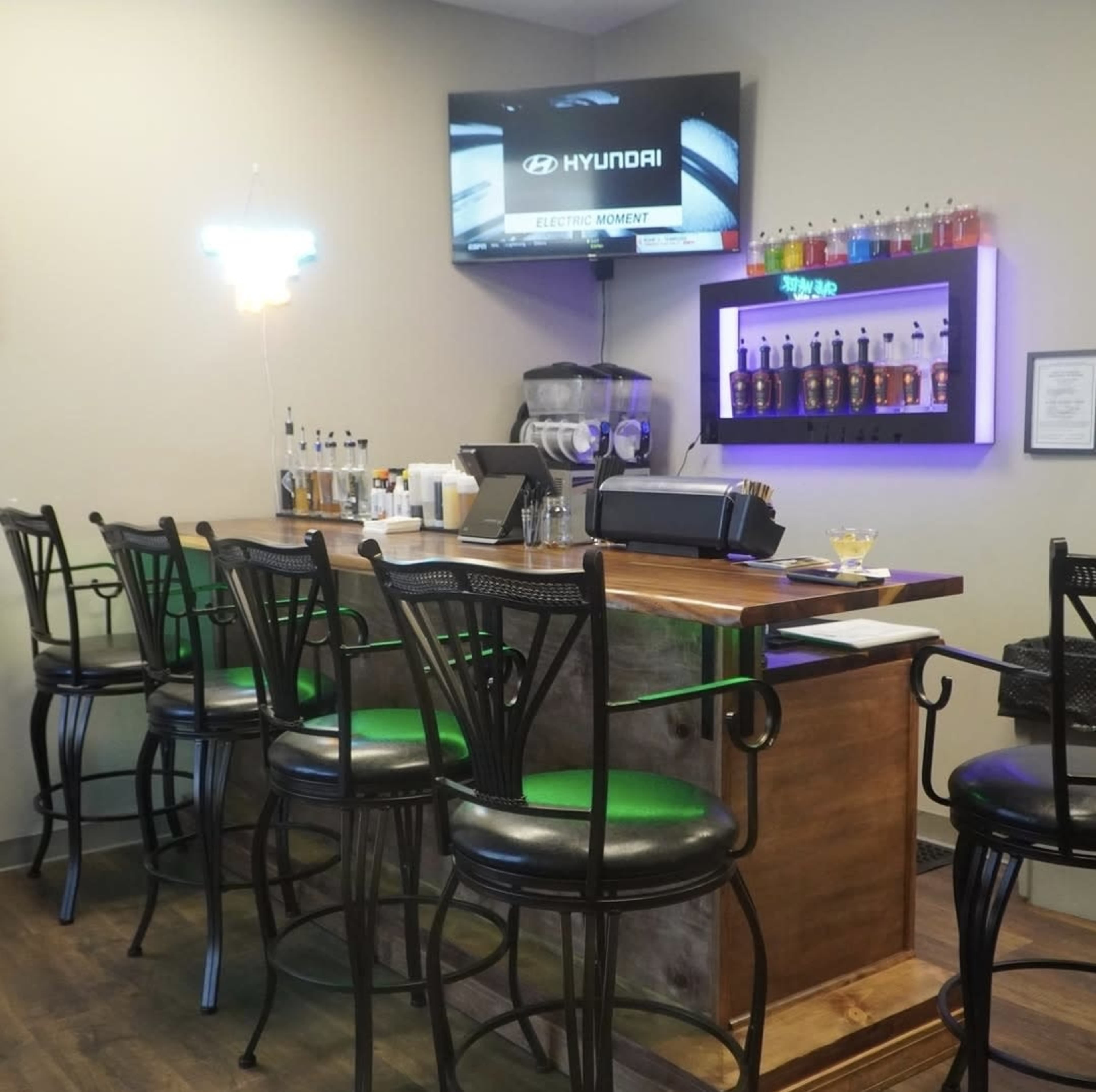 The image shows a modern bar with high stools, a wall-mounted television displaying a car advertisement, and colorful drinks arranged on a shelf behind the counter.