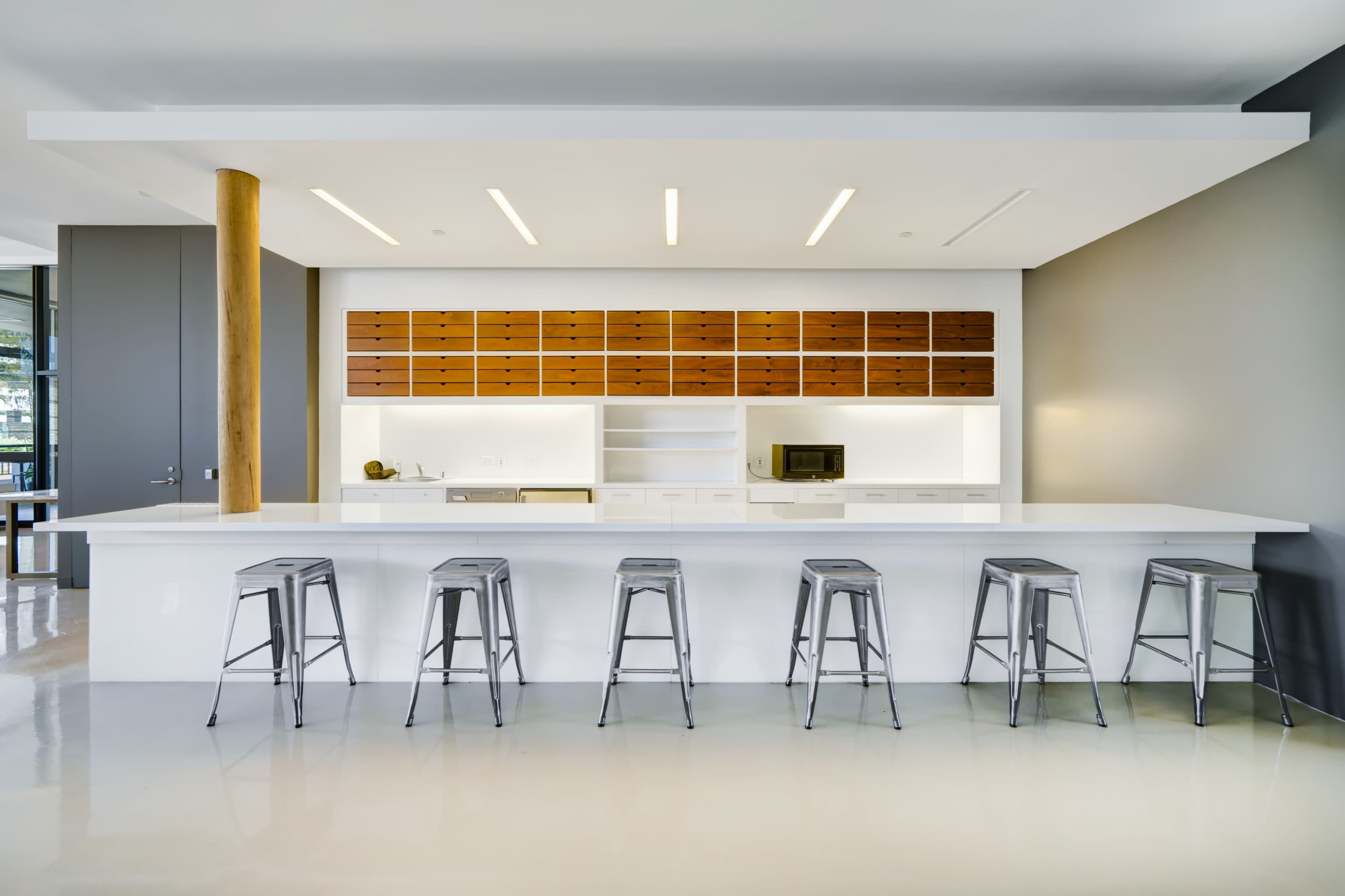 A modern kitchen features a white countertop with metal bar stools and a wooden cabinet overhead, illuminated by sleek linear lighting.