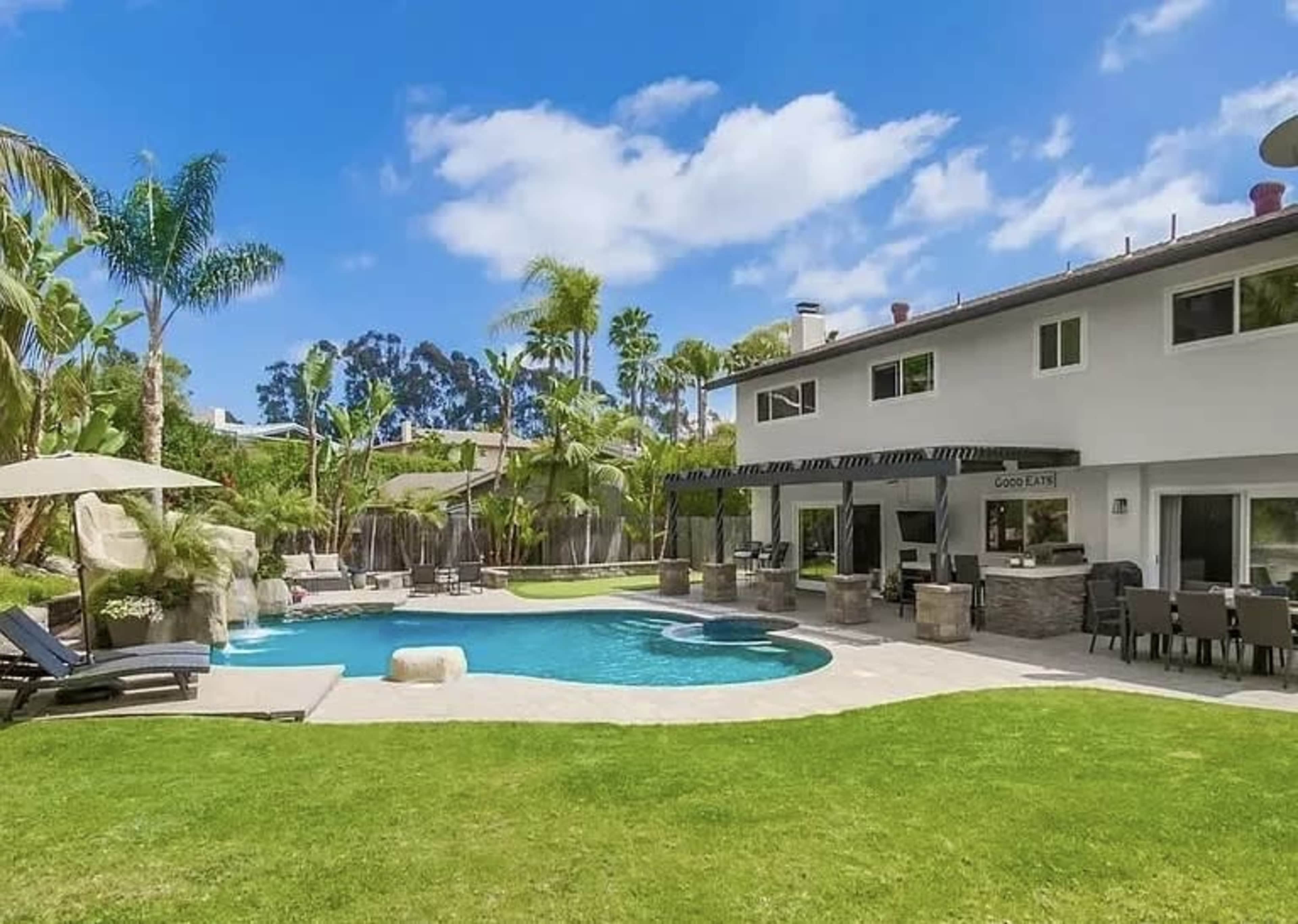 The image shows a spacious backyard with a swimming pool surrounded by palm trees, lounge chairs, and a two-story house.