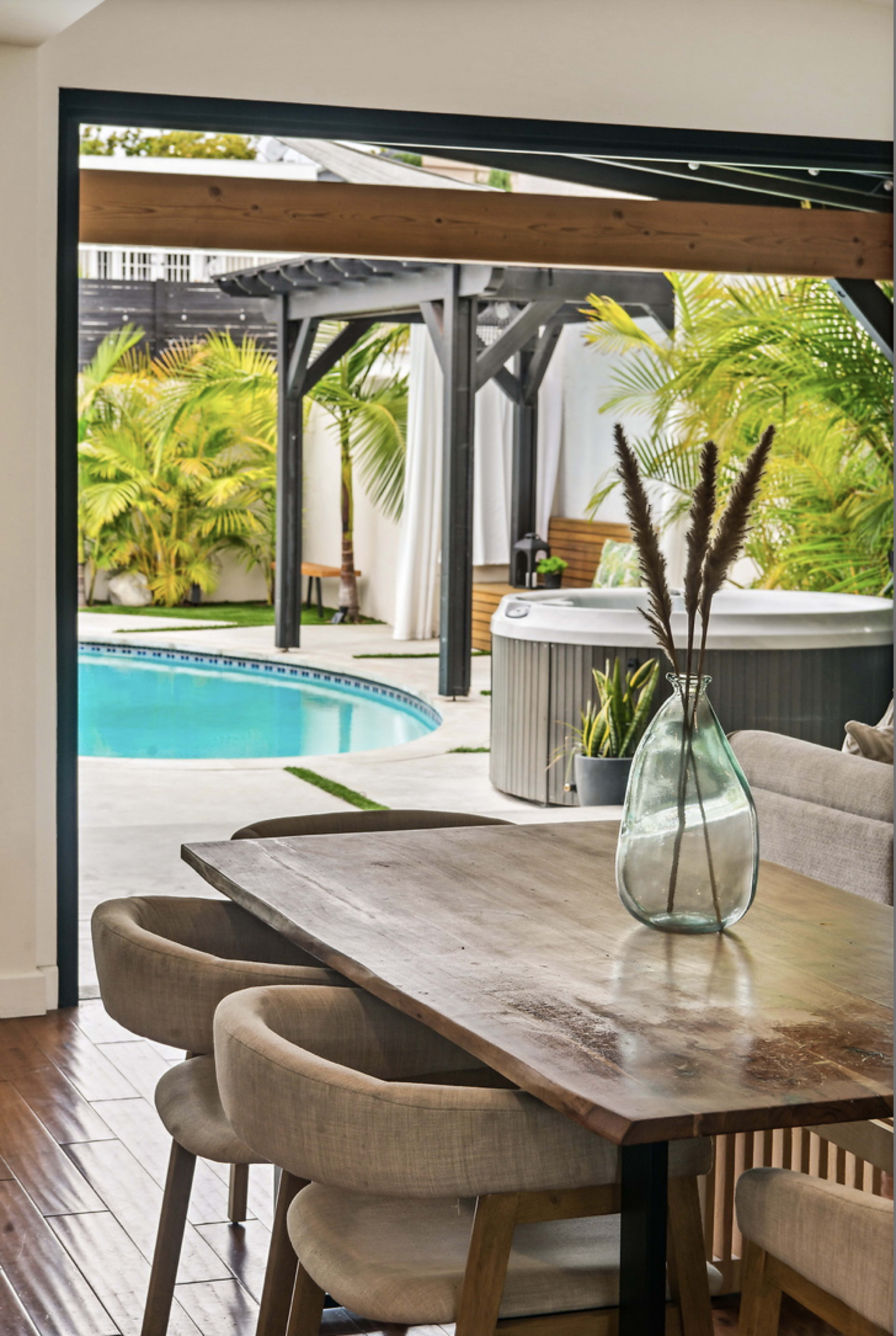 A wooden dining table with beige chairs is positioned indoors, overlooking a pool area surrounded by tropical plants and a hot tub.