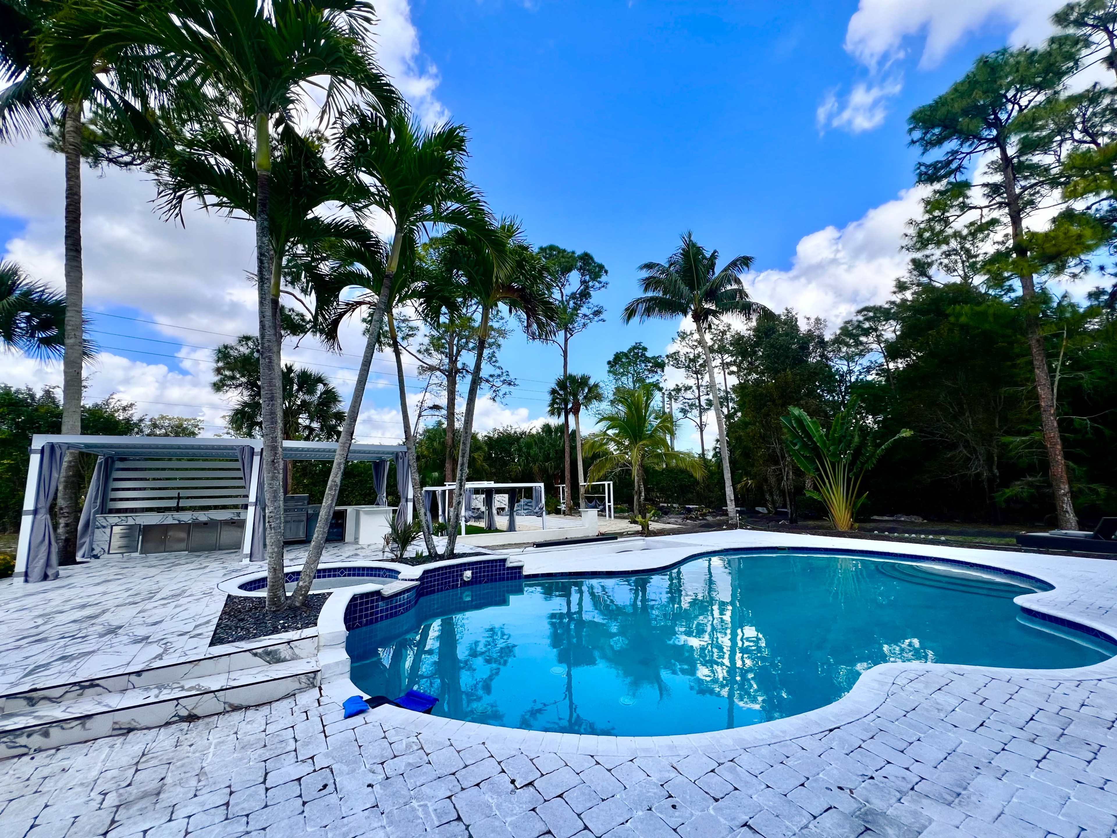 A sparkling blue pool is surrounded by palm trees and landscaped stone patios under a bright blue sky.