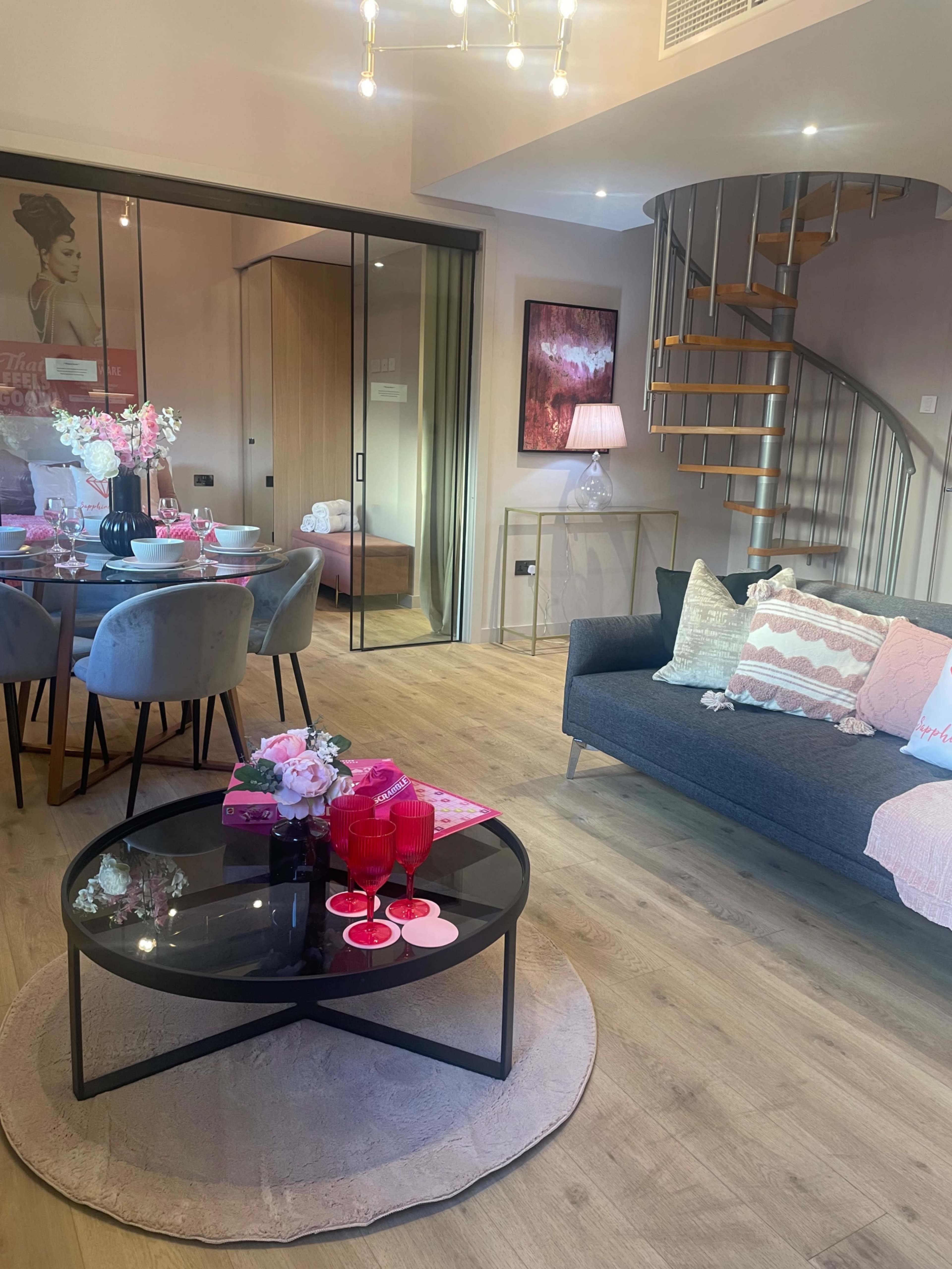 The image shows a modern living room with a spiral staircase, a round coffee table, a dining area, and pink decor elements.