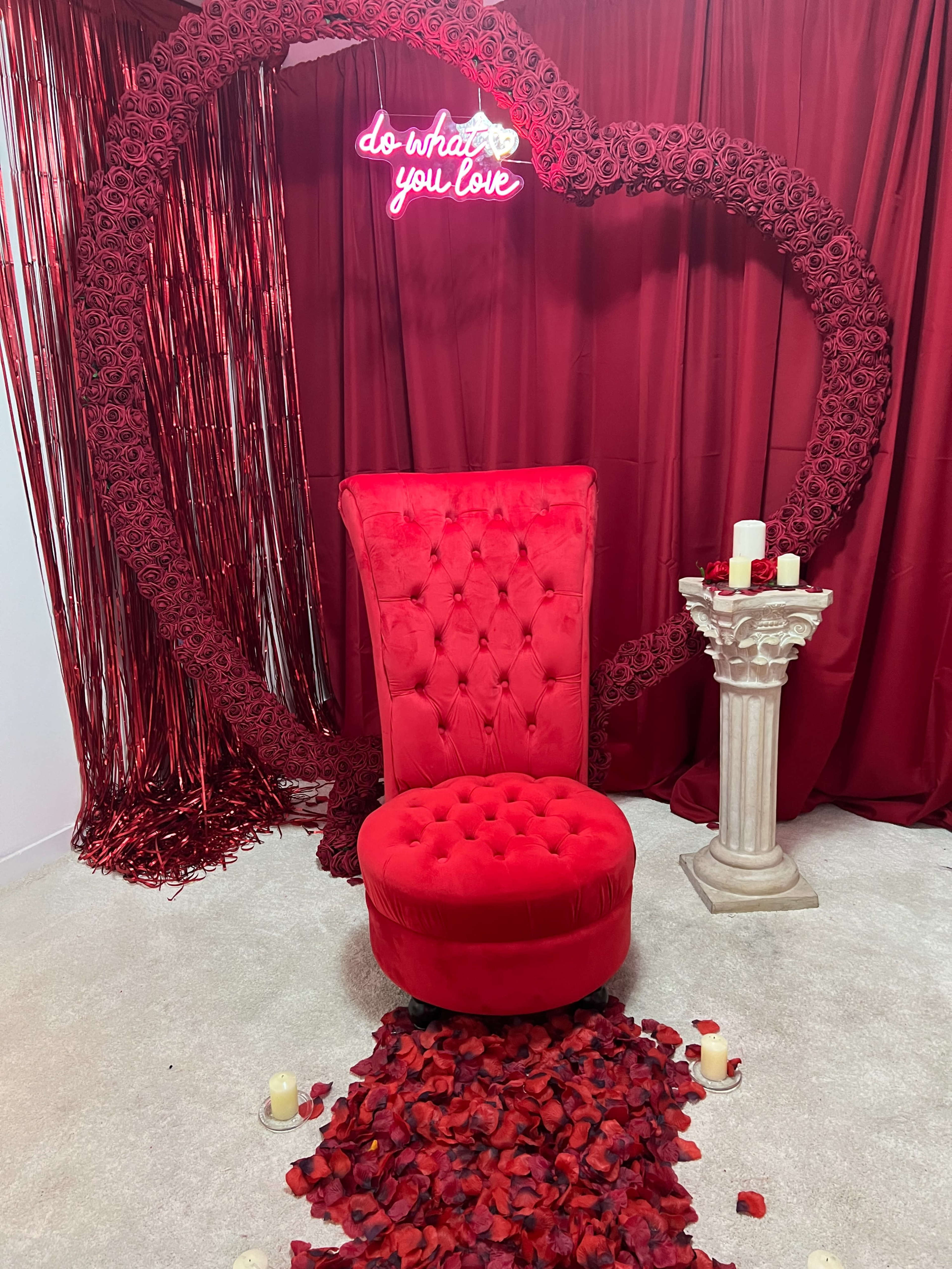 A red velvet chair is positioned in front of a heart-shaped arrangement of roses, with a neon sign reading "do what you love" in the background.