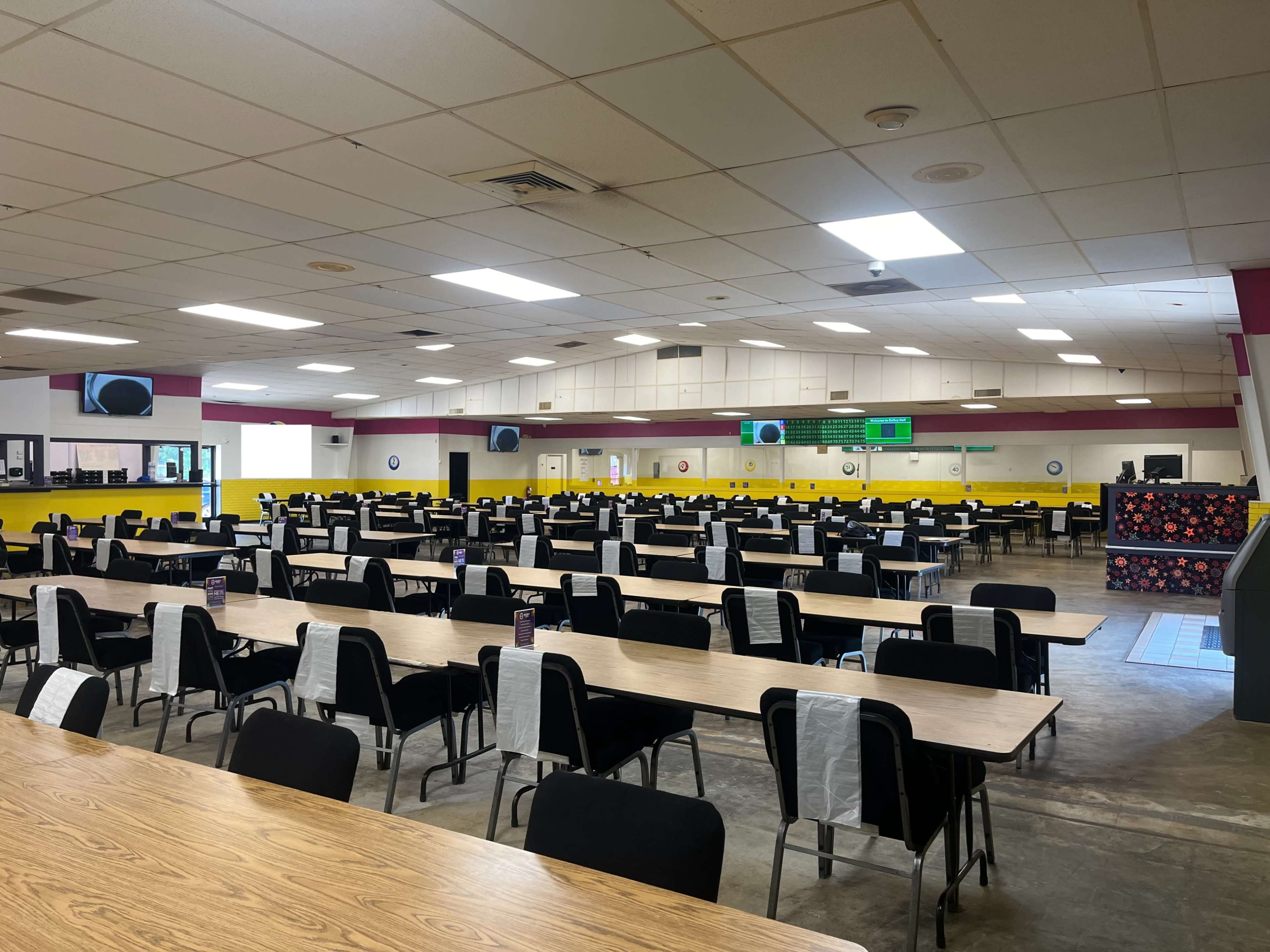 A large cafeteria with numerous empty tables and chairs arranged in rows, illuminated by overhead lights.