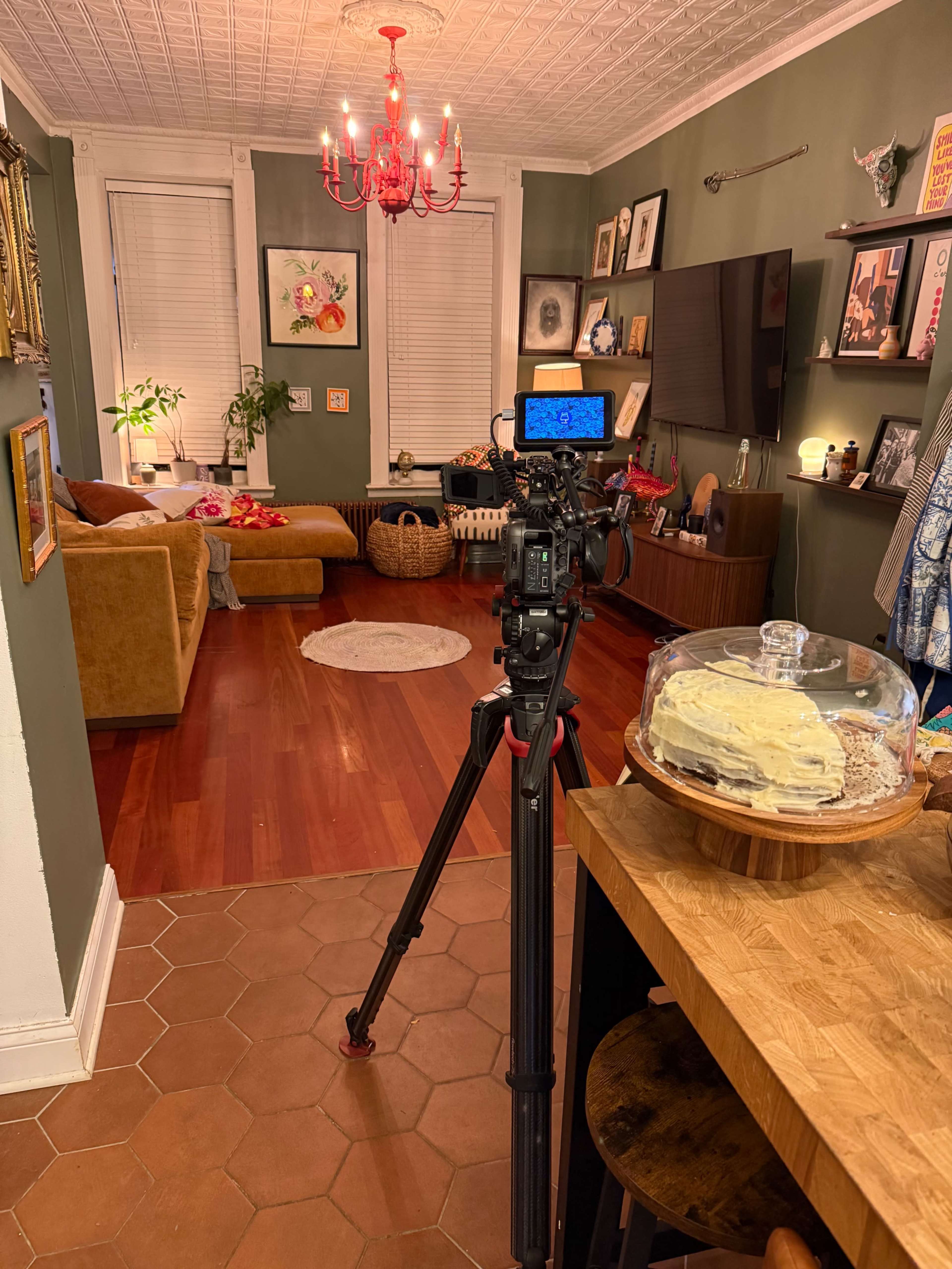 A camera on a tripod is set up in a well-decorated living room with a chandelier, a couch, and a table displaying a cake under a glass dome.