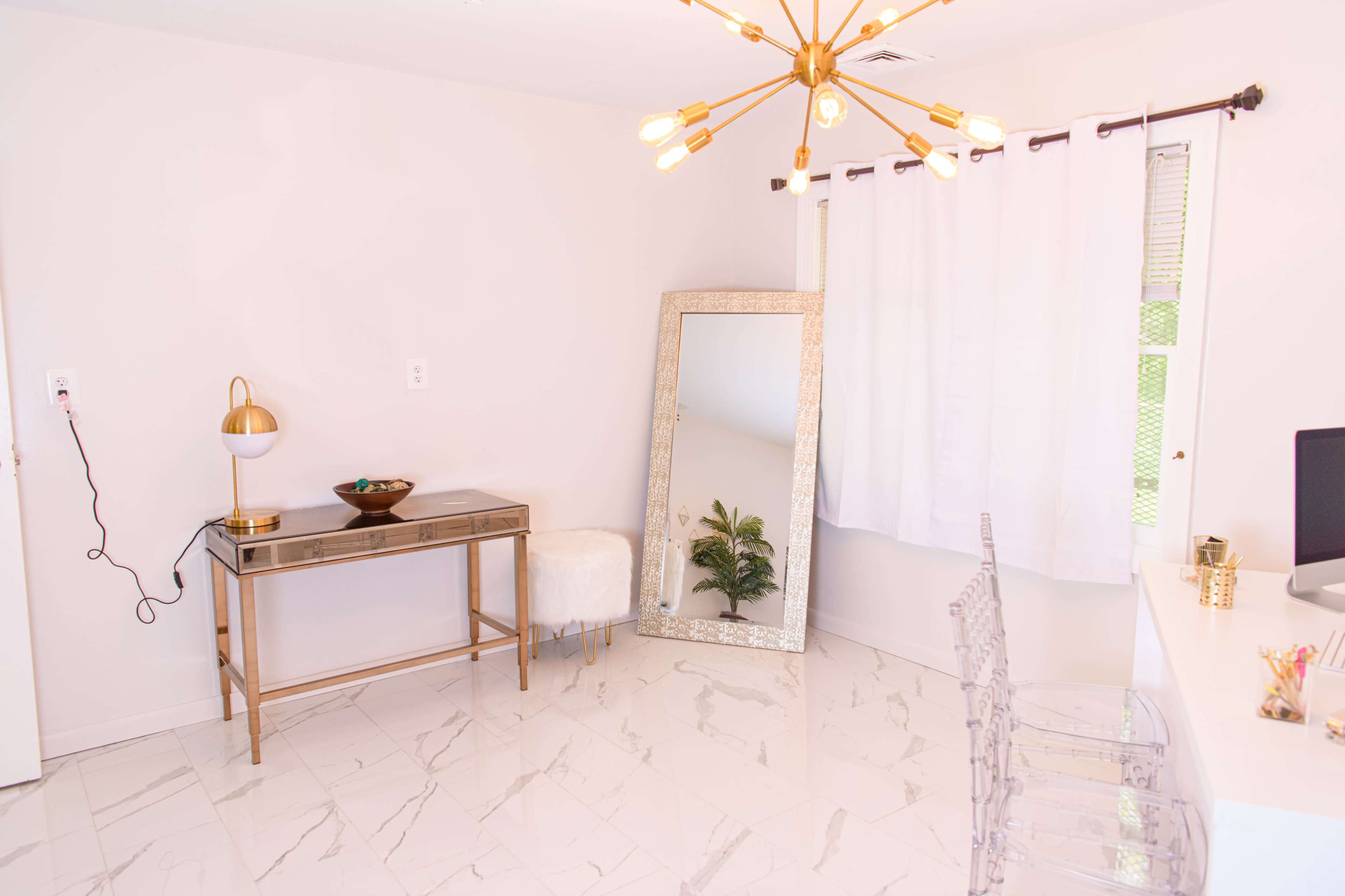 A modern room features a gold-accented vanity table, a large mirrored frame, a lamp, and clear acrylic chairs against a backdrop of white walls and a window with sheer curtains.