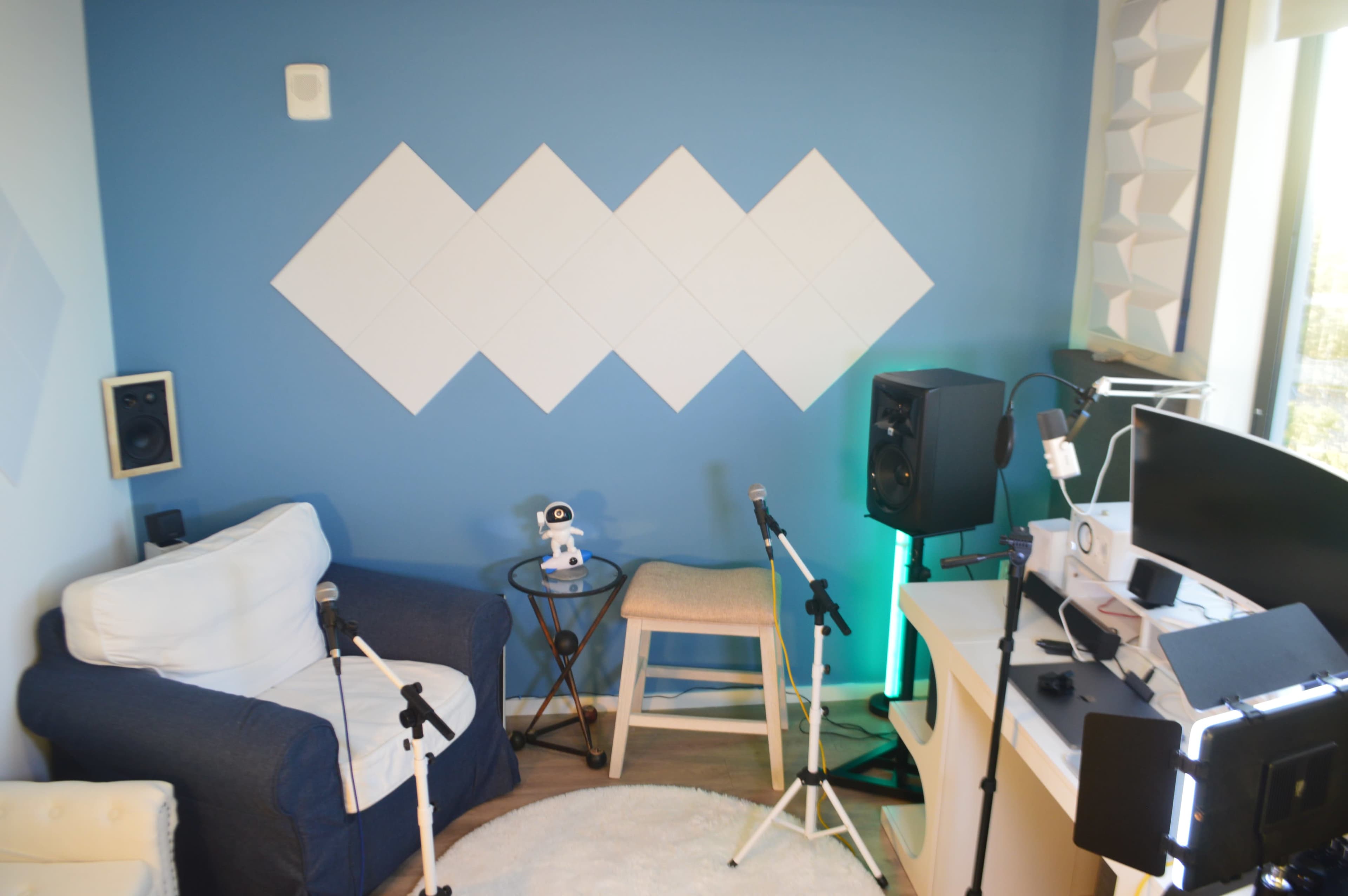 The image shows a small recording studio setup featuring a blue wall with diamond-shaped panels, a white couch, a stool, two microphones on stands, and various audio equipment on a desk.