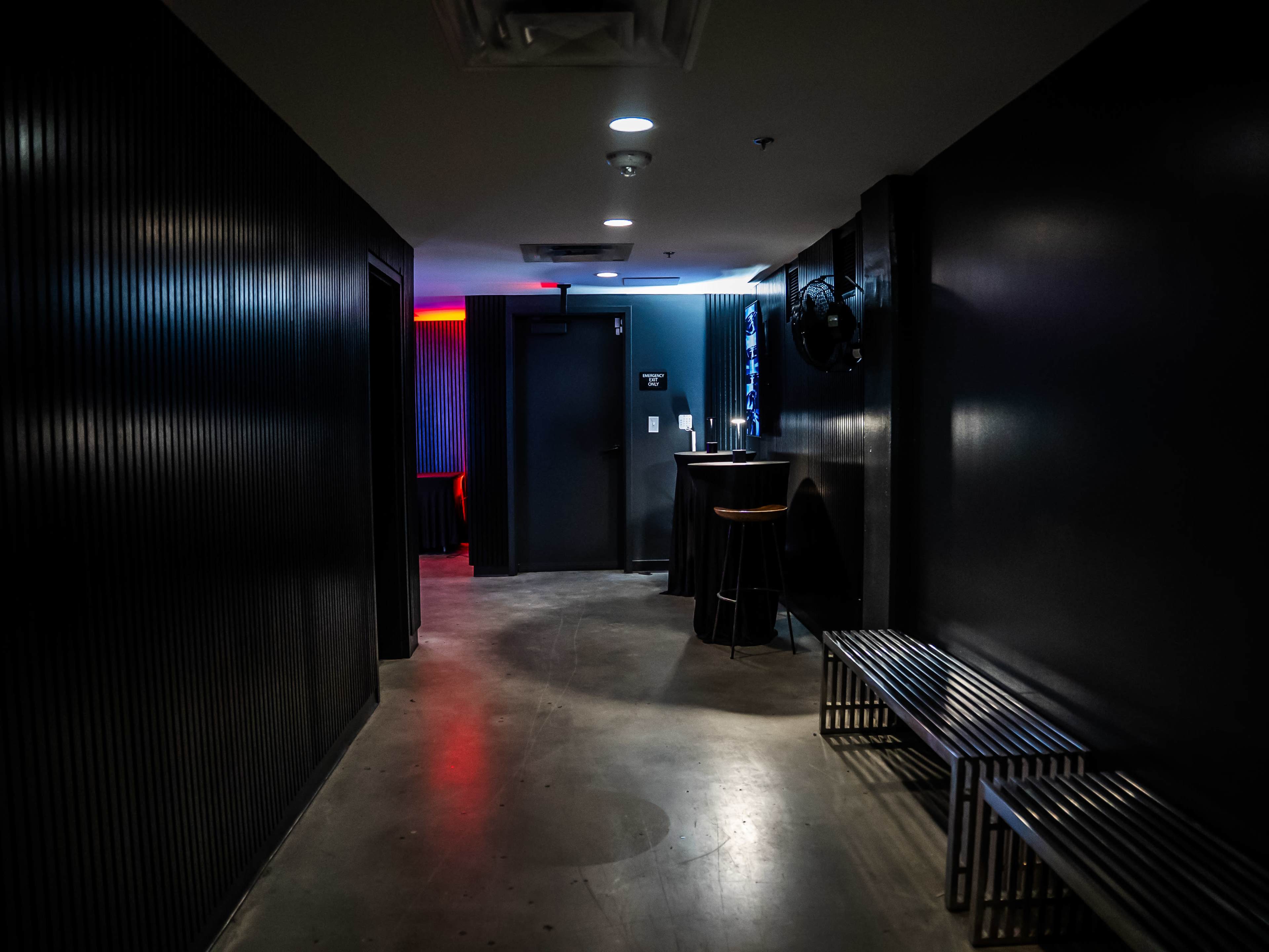The image shows a dimly lit hallway with black walls, a door at the end, and metal benches along the sides.