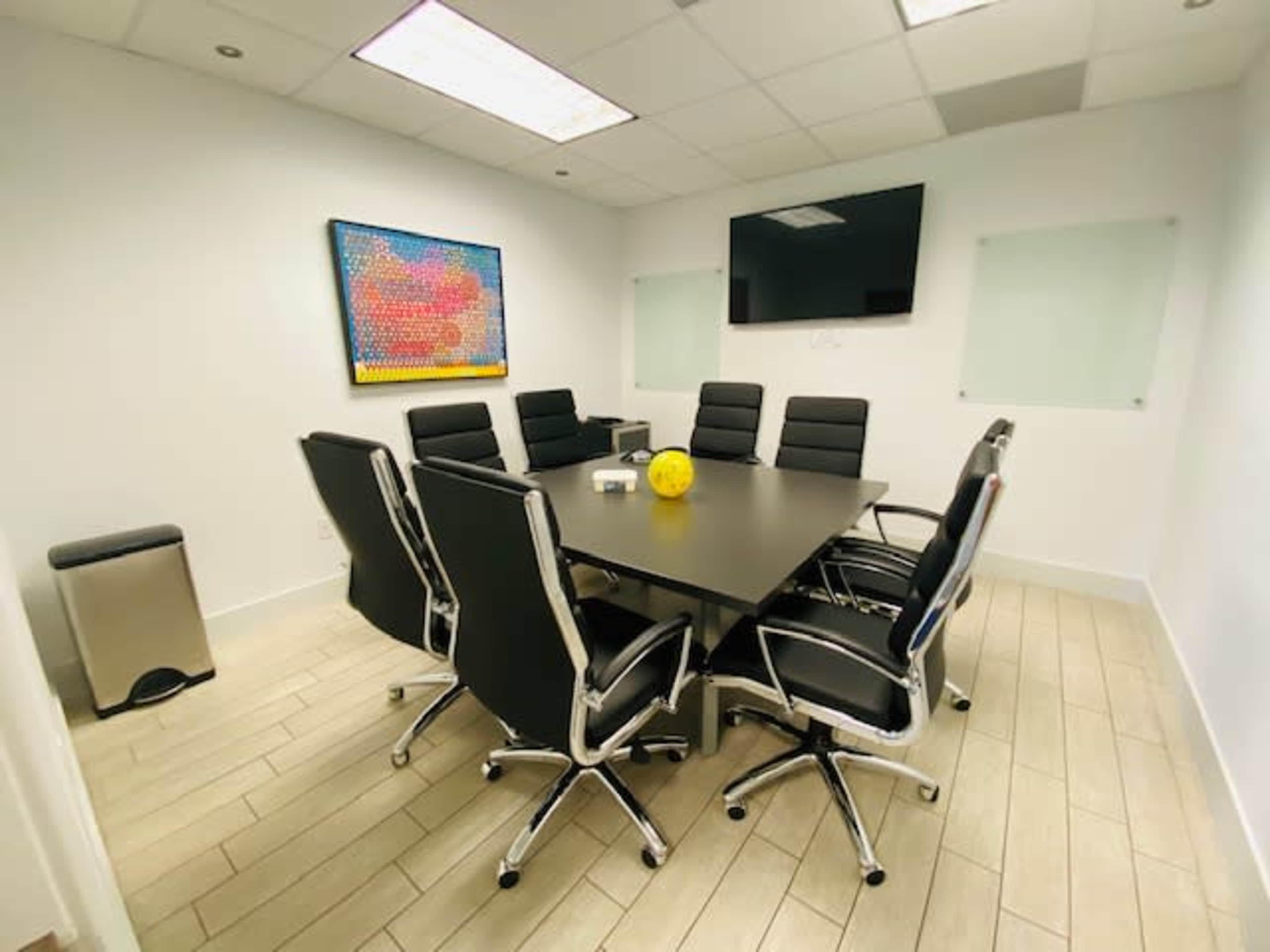A modern conference room features a long table surrounded by black ergonomic chairs, with a large screen and colorful artwork on the wall.