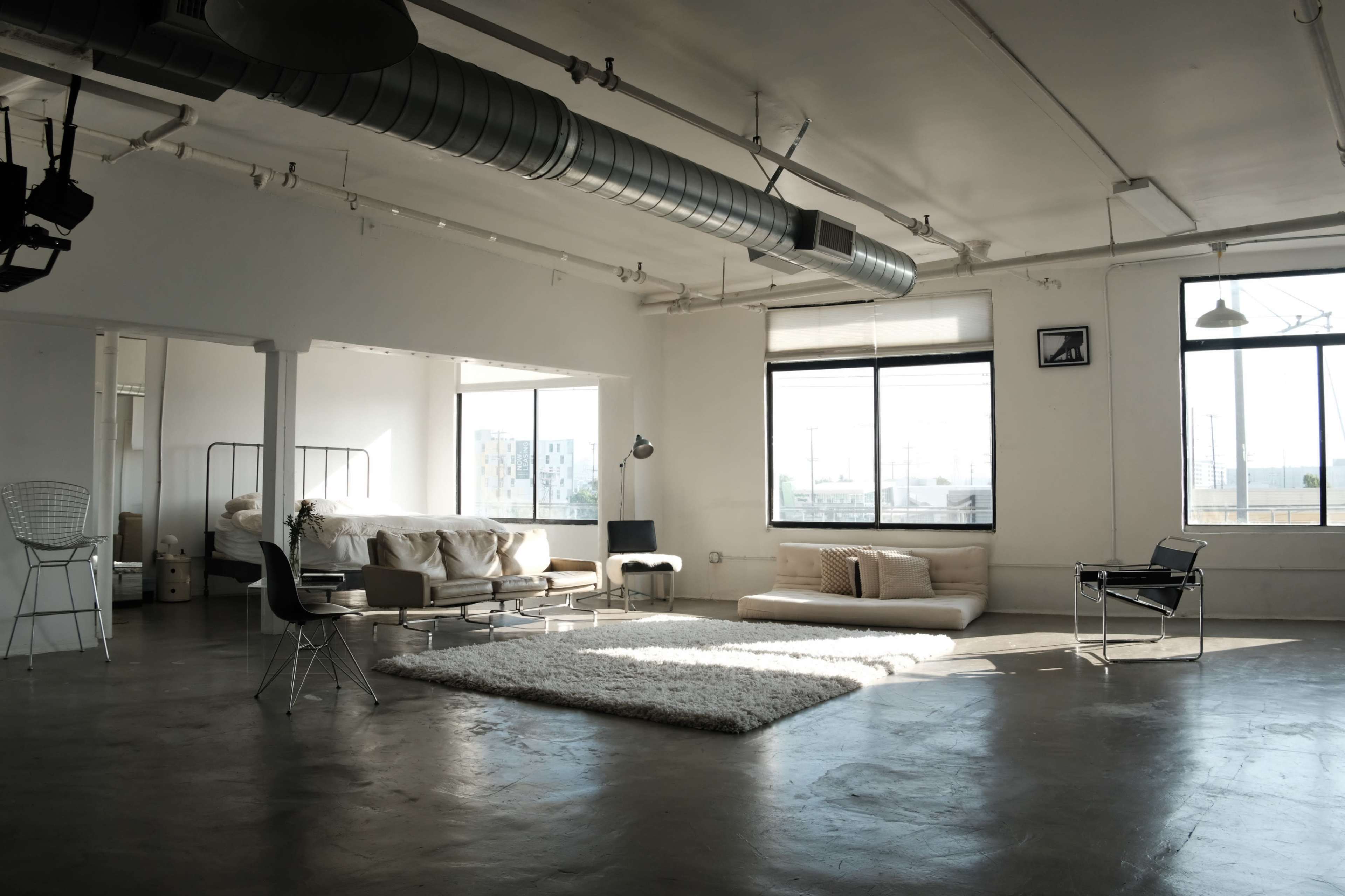 The image shows a spacious, modern loft with concrete flooring, large windows, and minimalist furniture, including a sofa, bed, and rugs.
