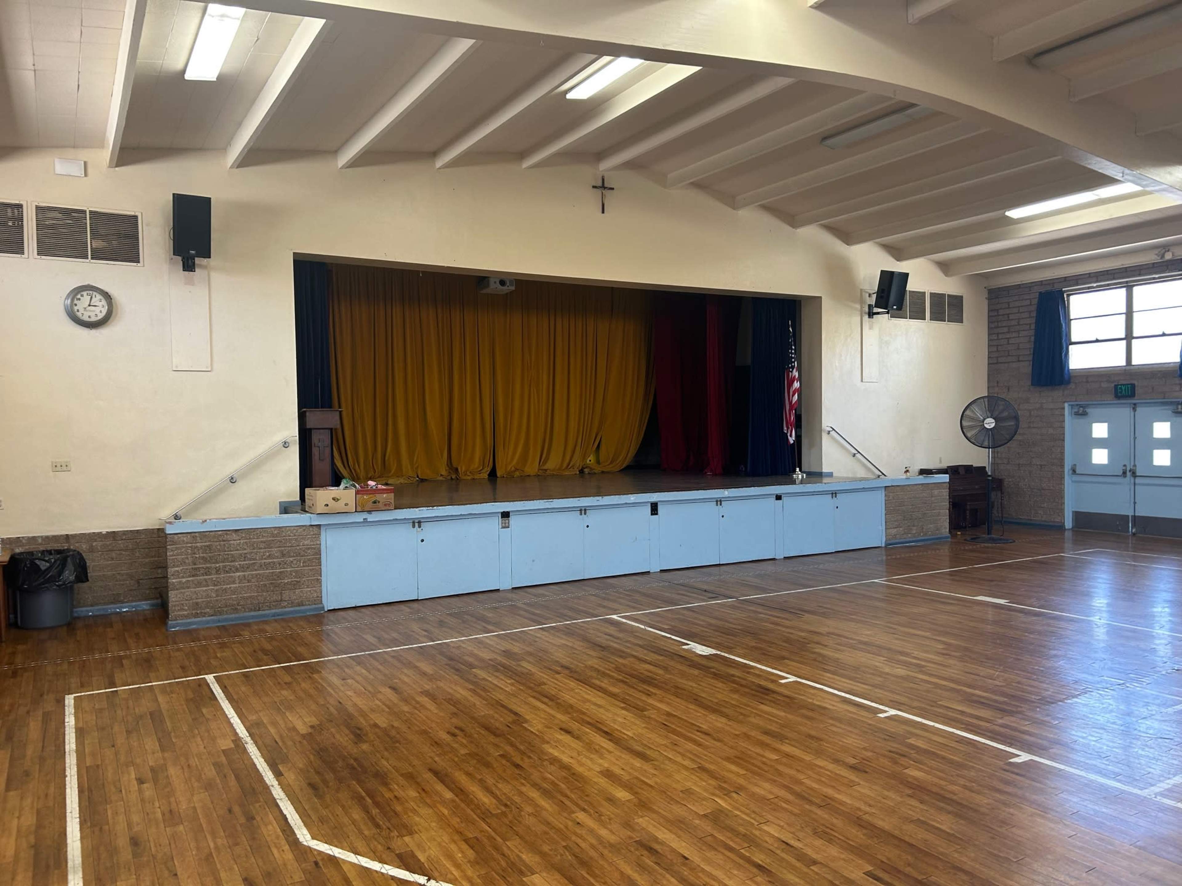 The image shows an empty gymnasium with a stage featuring yellow and red curtains, an American flag, and wooden flooring.
