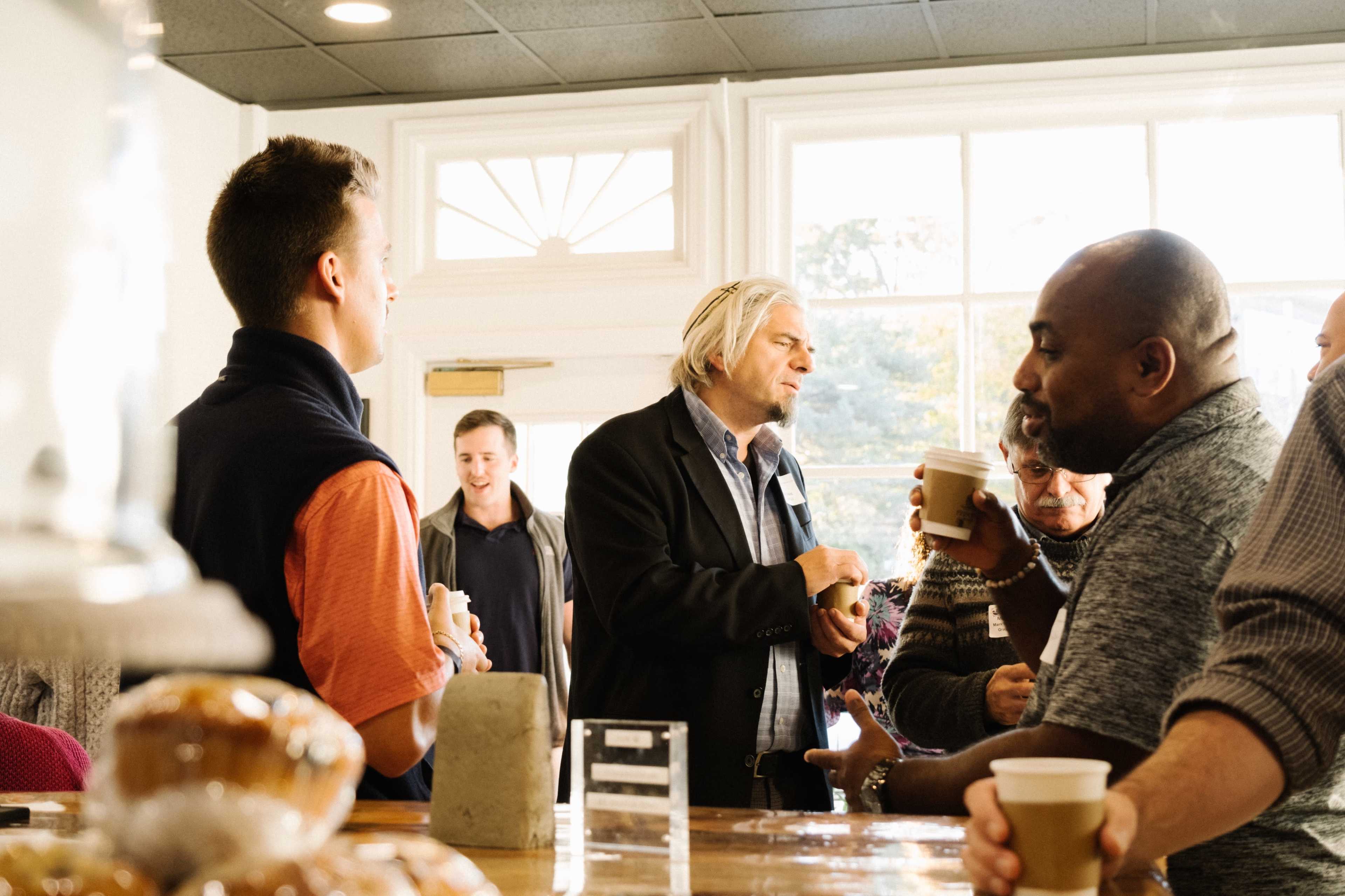 A group of people are engaging in conversation inside a well-lit café, with some holding coffee cups and pastries on display nearby.