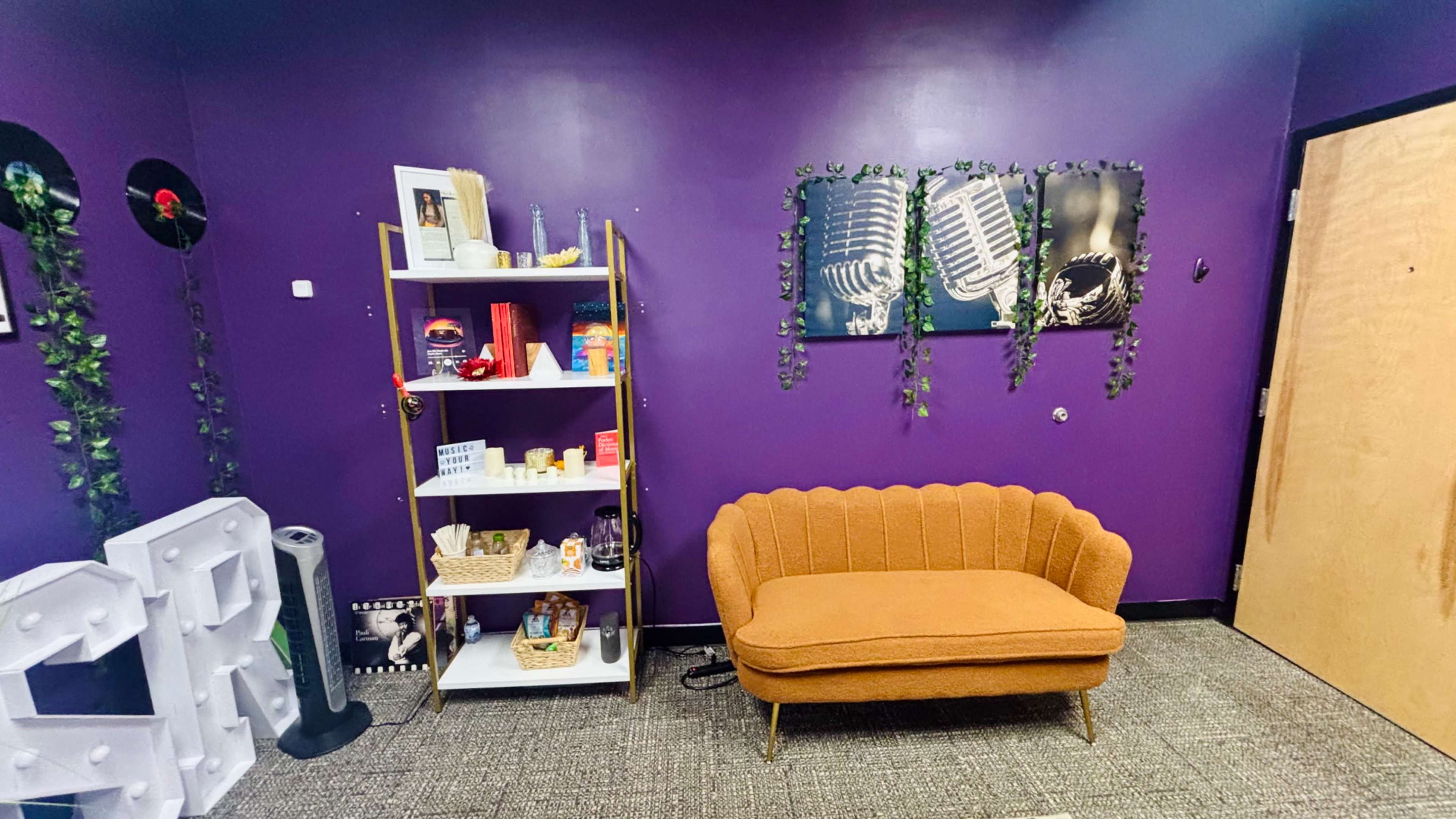 The image shows a small room with purple walls, featuring an orange couch, a bookshelf filled with various items, and framed art of microphones.