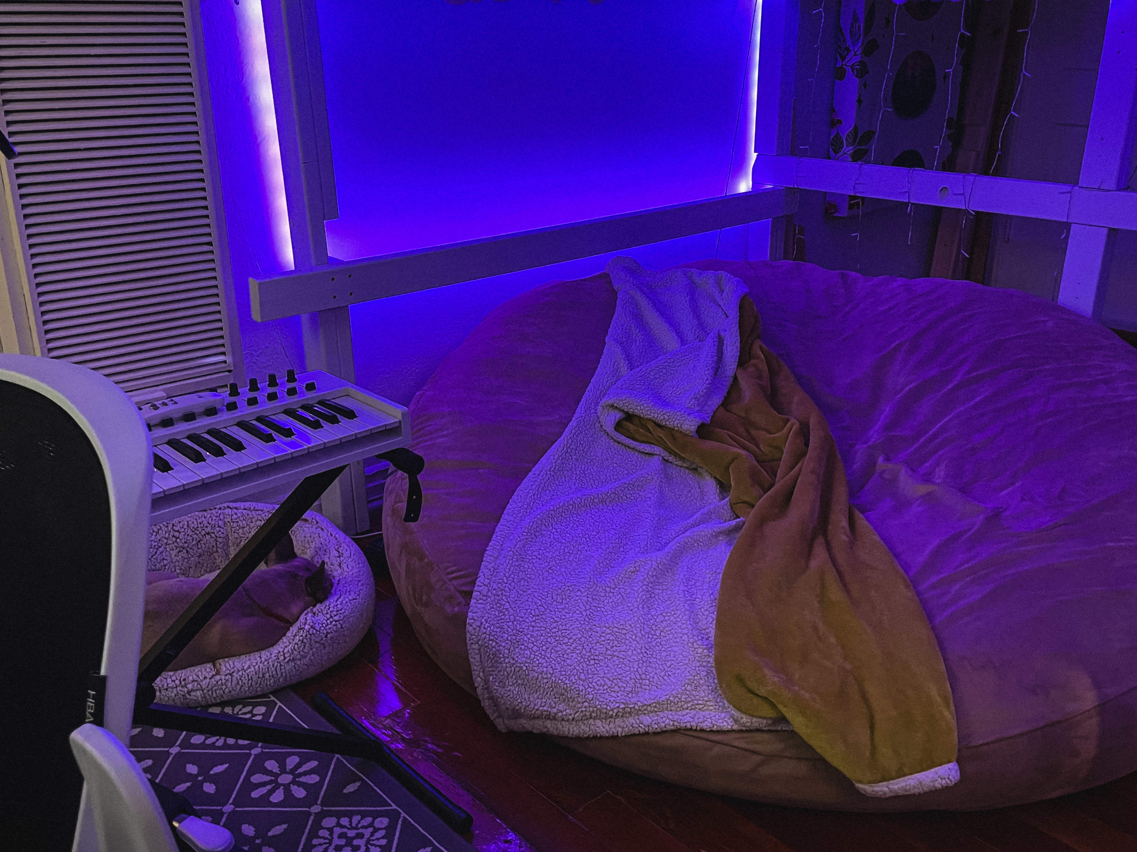 A room features a large bean bag chair with a blanket draped over it, alongside a keyboard on a stand and ambient purple lighting.