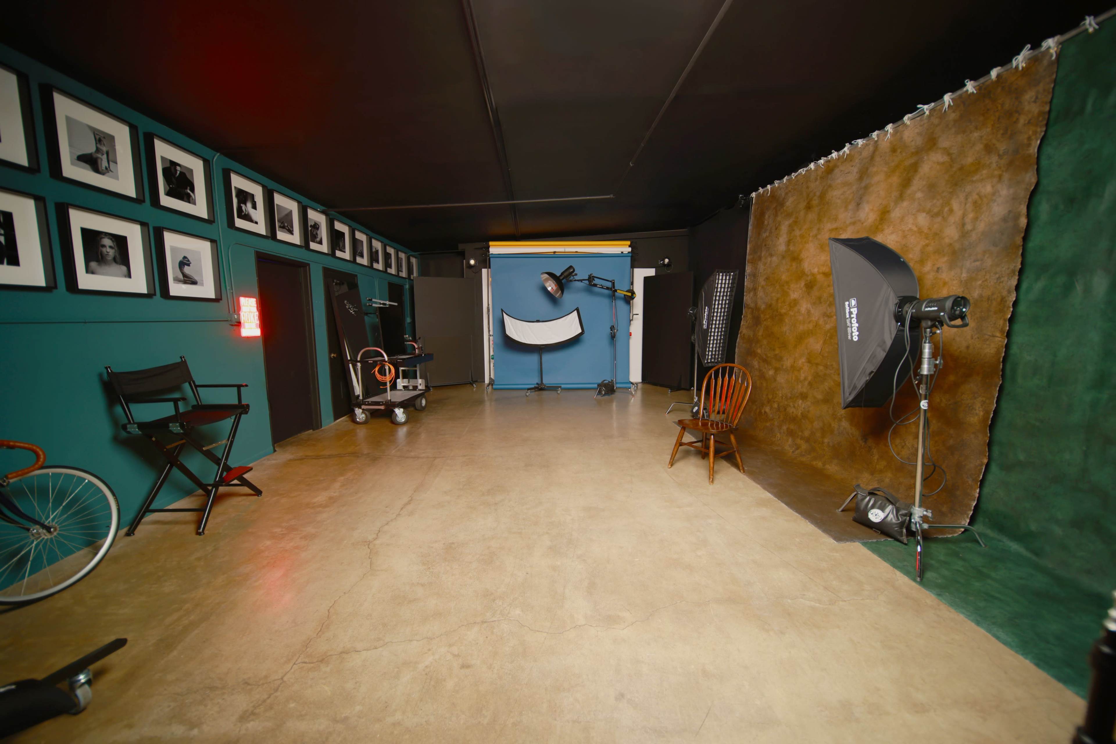 The image shows an empty photographic studio with various lighting equipment, a backdrop, and framed black-and-white photographs on the walls.