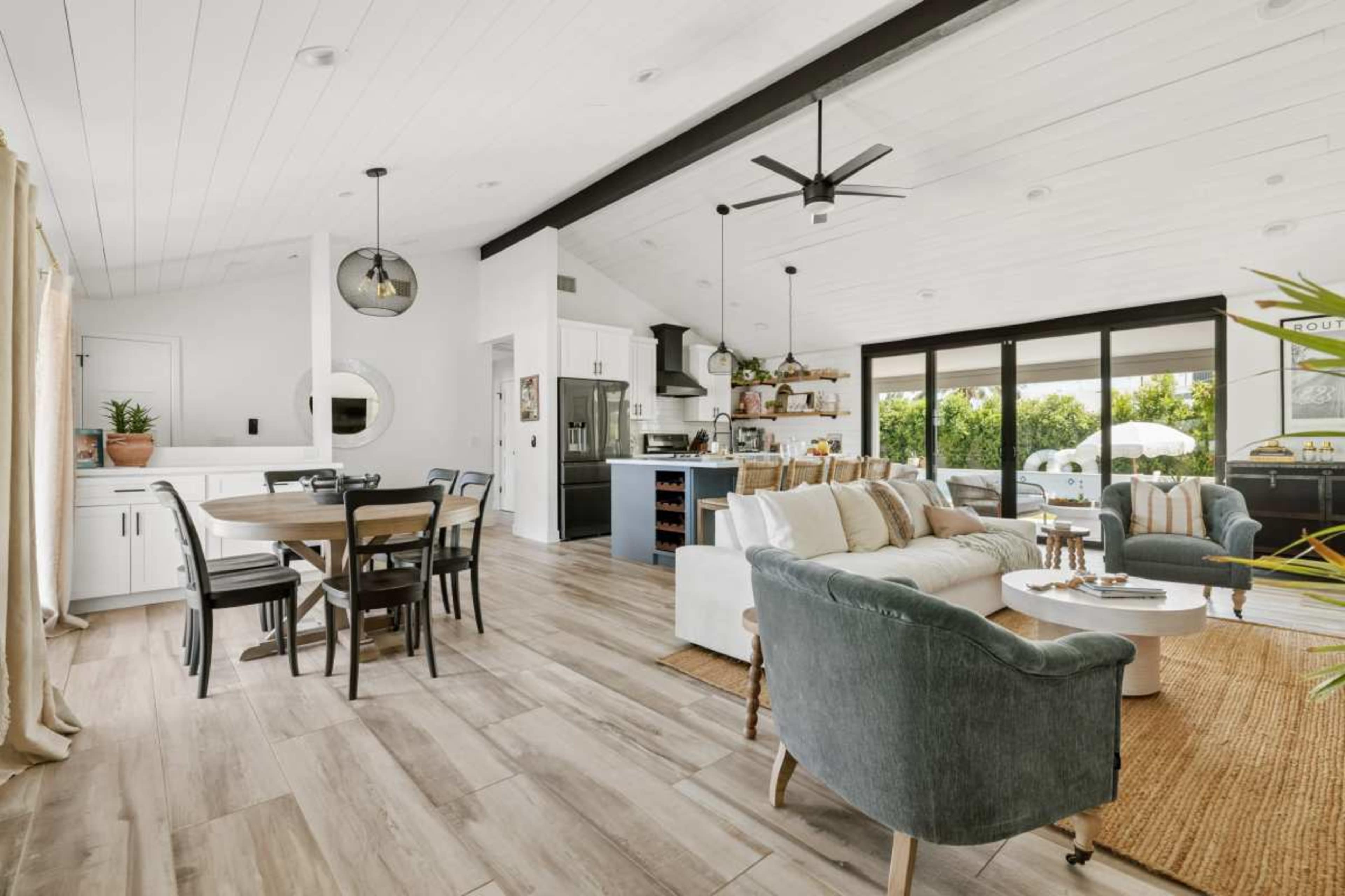 The image shows a modern open-concept living space featuring a white sofa, a grey armchair, a dining table, and large windows that provide ample natural light.