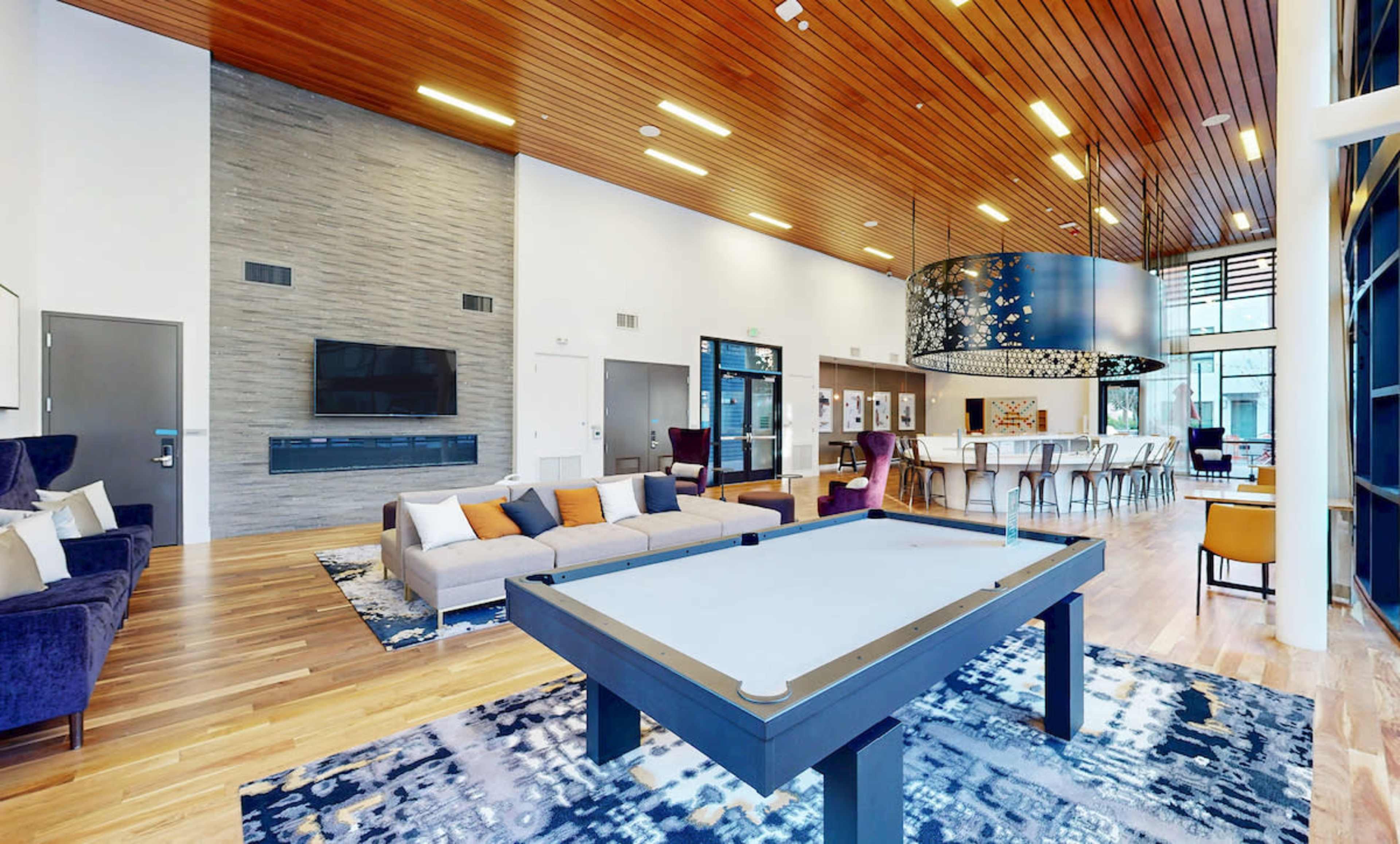 The image shows a modern lounge area featuring a pool table, a large sectional sofa, and a dining table under a decorative ceiling.