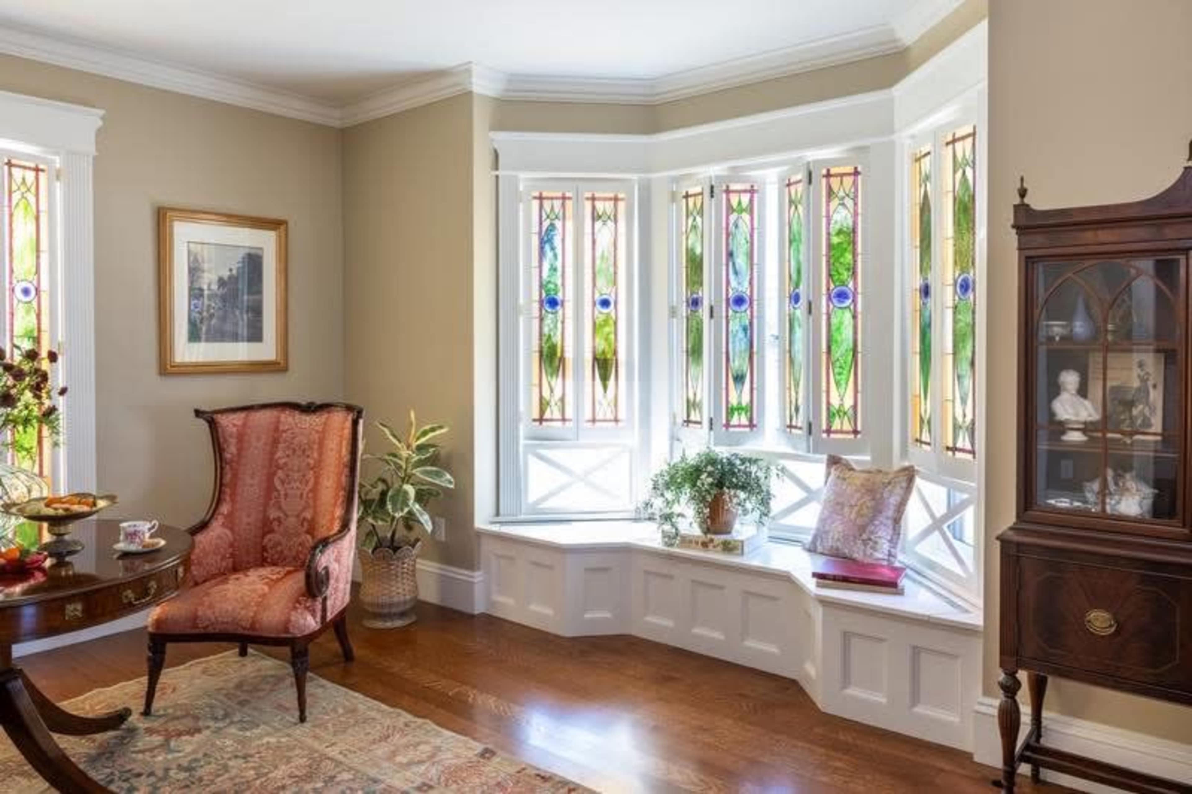 The image shows a brightly lit living room featuring a window bay with stained glass, an ornate chair, a small table with decorative items, and a wooden cabinet.