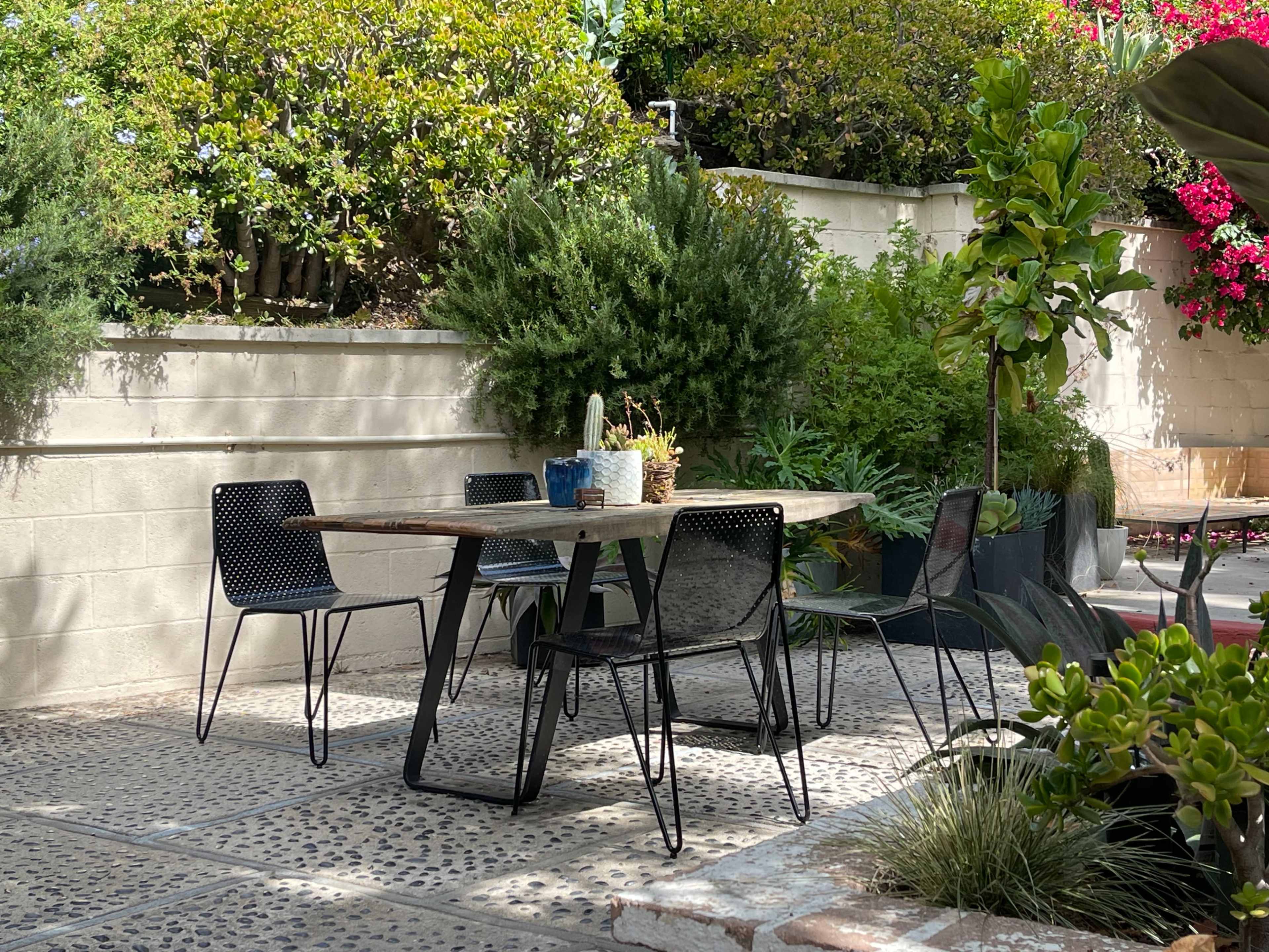 A rectangular wooden table with four black mesh chairs is positioned in a patio surrounded by greenery and potted plants.