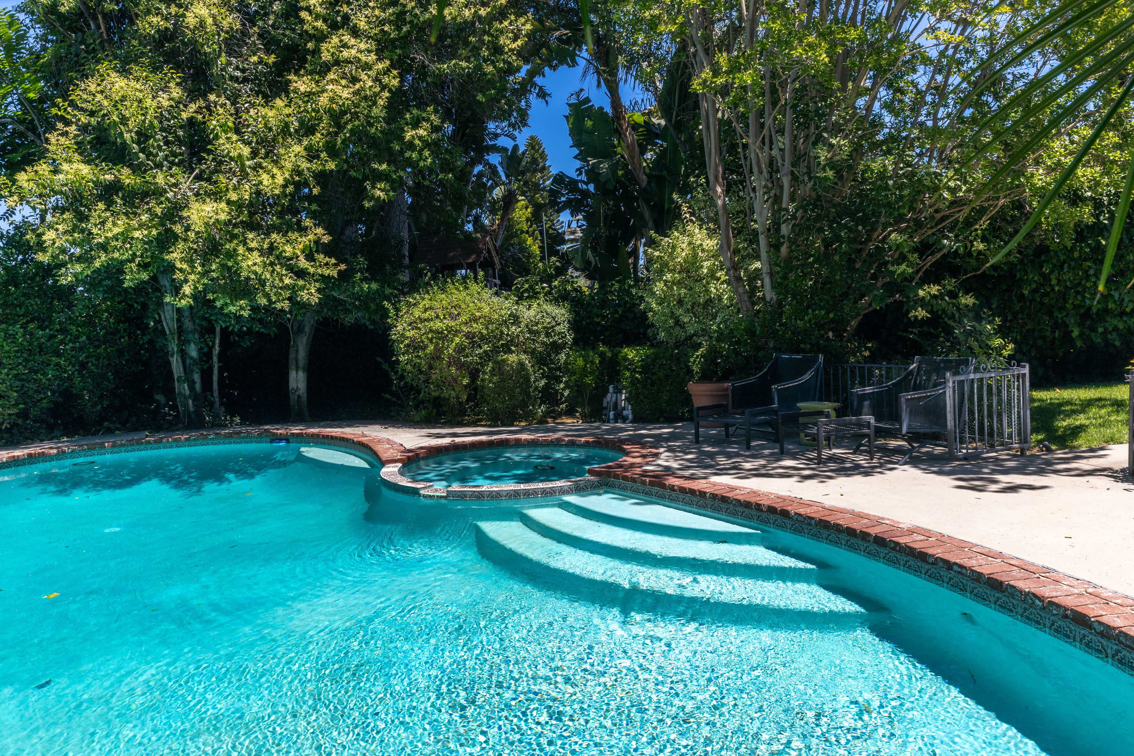 The image shows a swimming pool with a connected hot tub, surrounded by lush greenery and patio furniture.