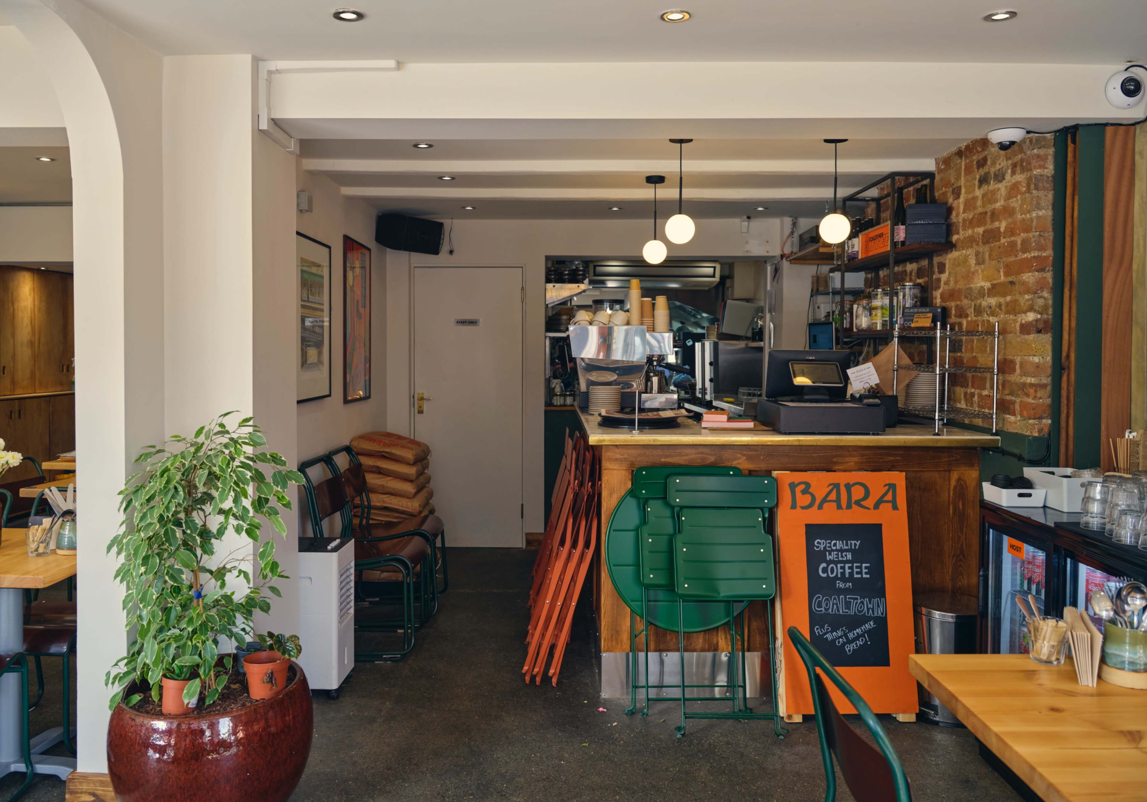 The image shows a modern café interior with a wooden counter, stacked chairs, and a potted plant in a corner.
