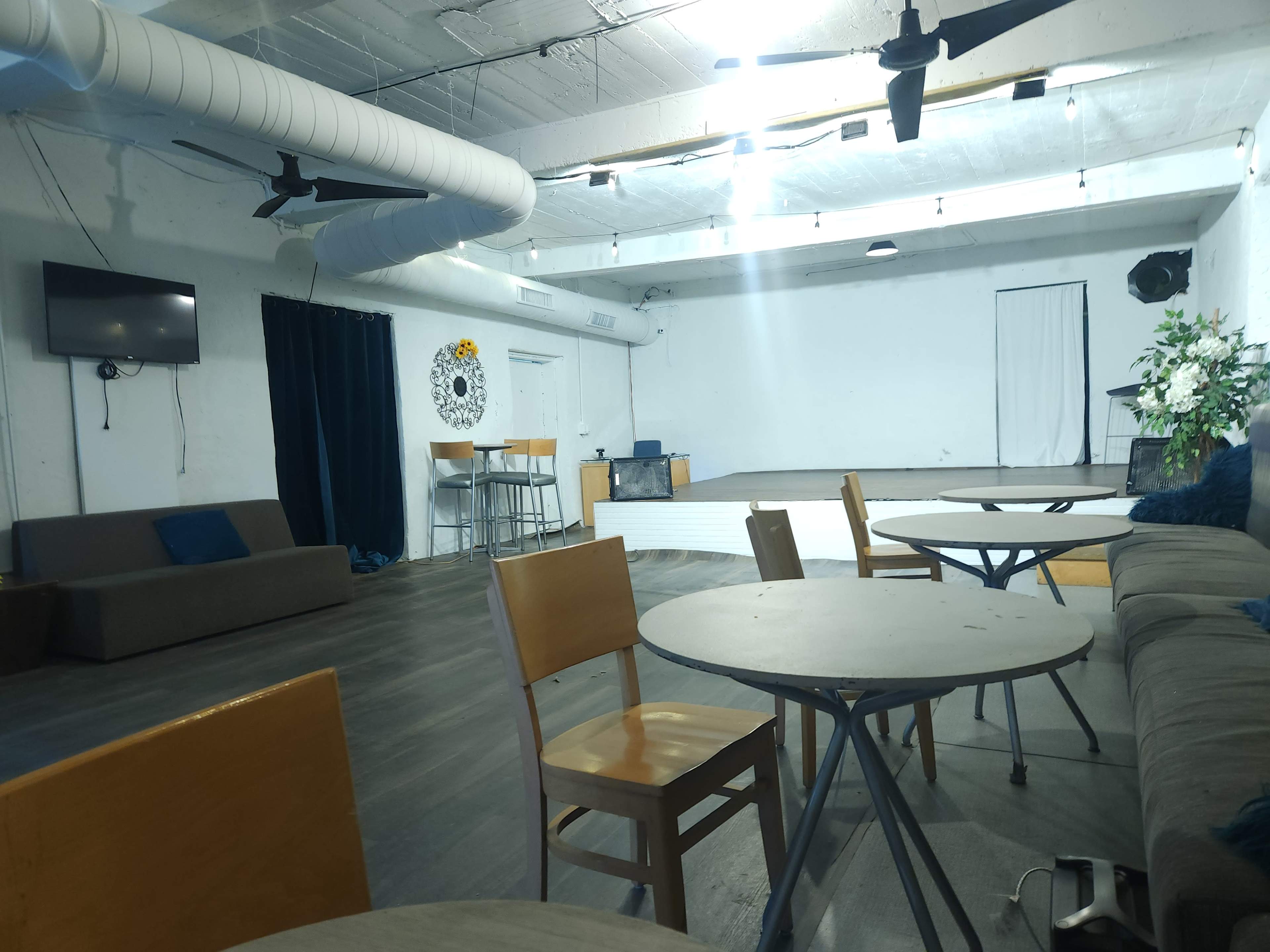 An empty event space with several tables and chairs arranged in front of a couch, while a bar area is positioned in the background.