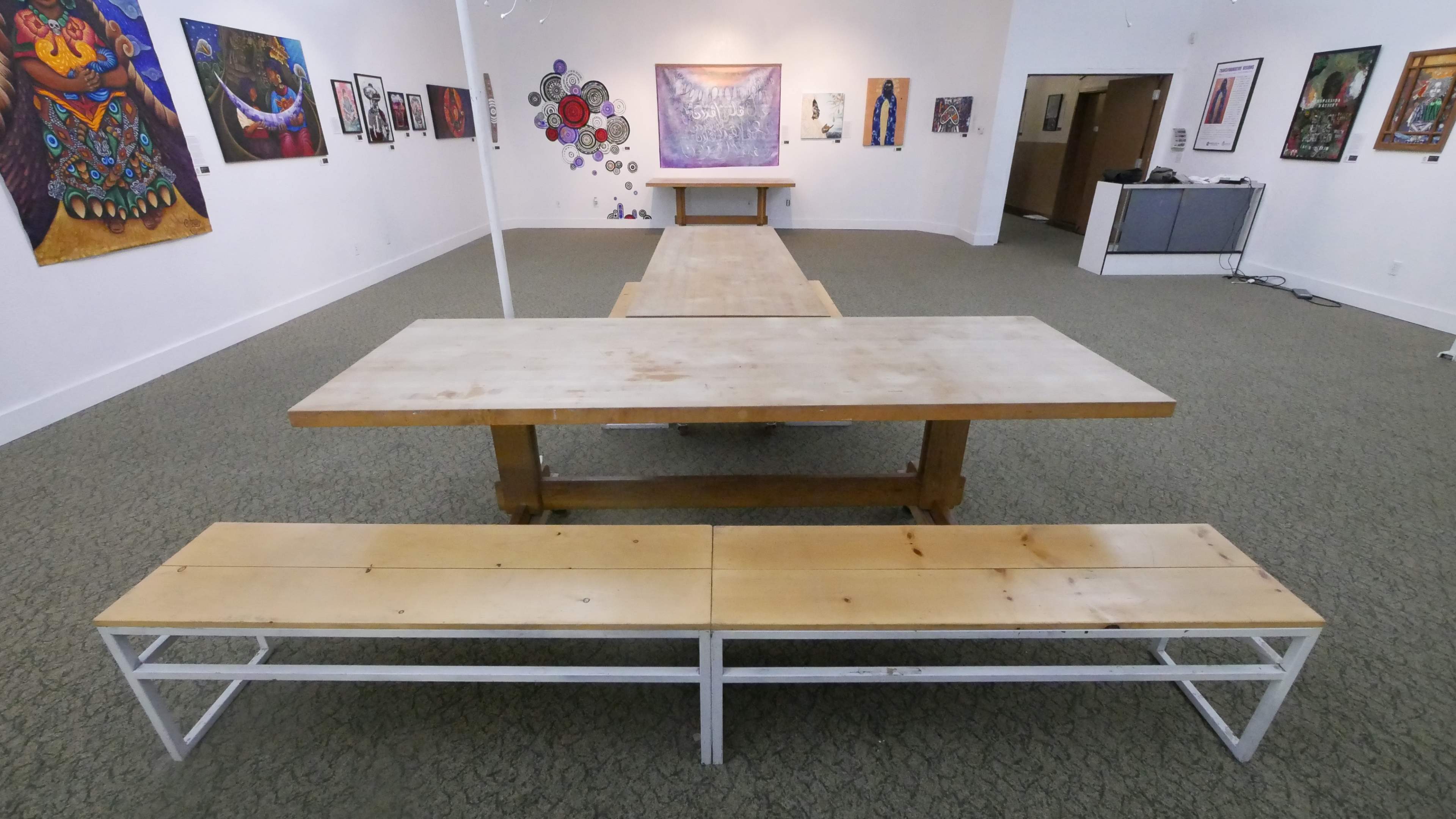 A spacious art gallery featuring a long wooden table with benches, surrounded by colorful artwork displayed on the walls.