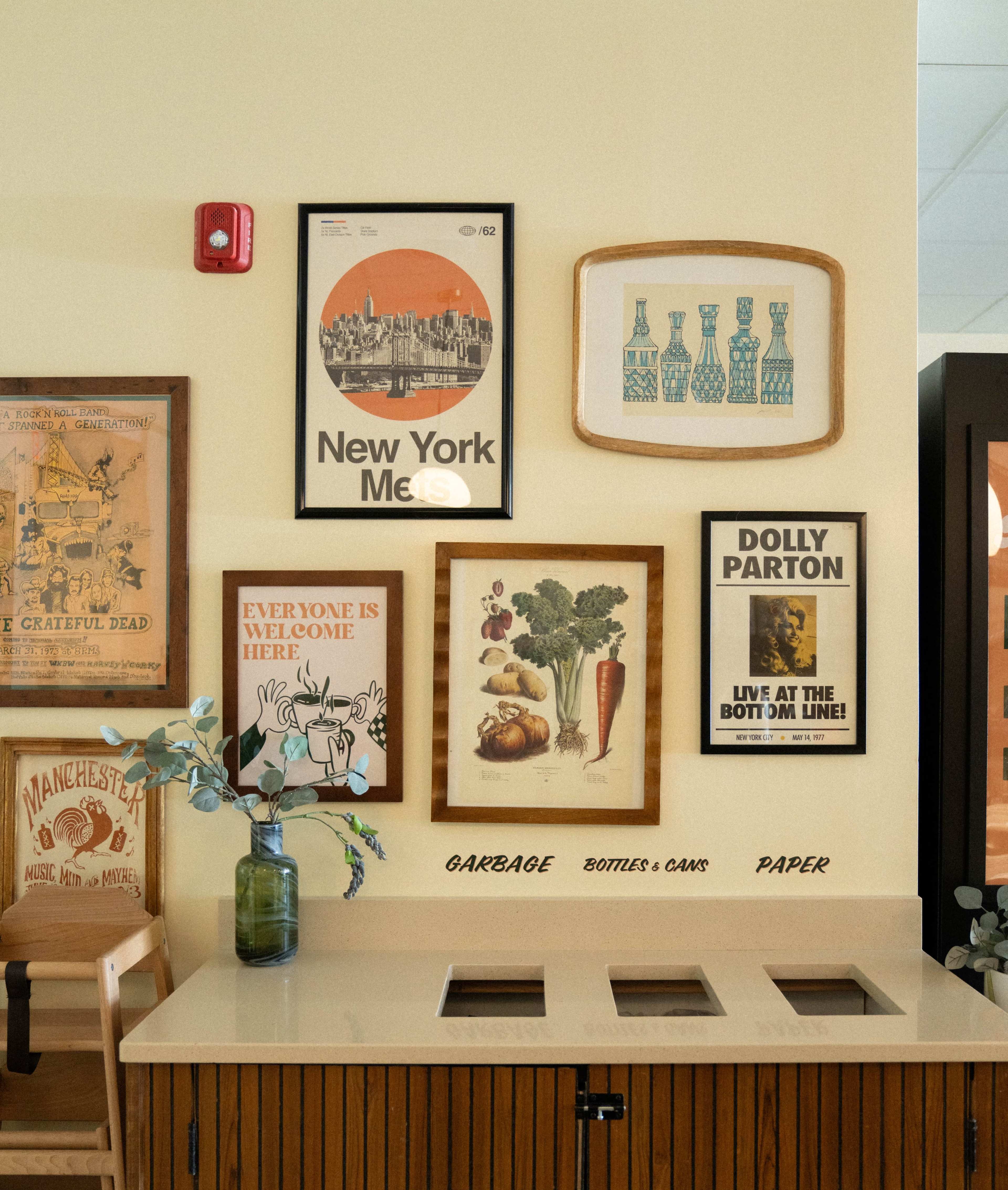 The image shows a decorated wall featuring various framed artworks and posters above a recycling and waste station labeled for garbage, bottles & cans, and paper.