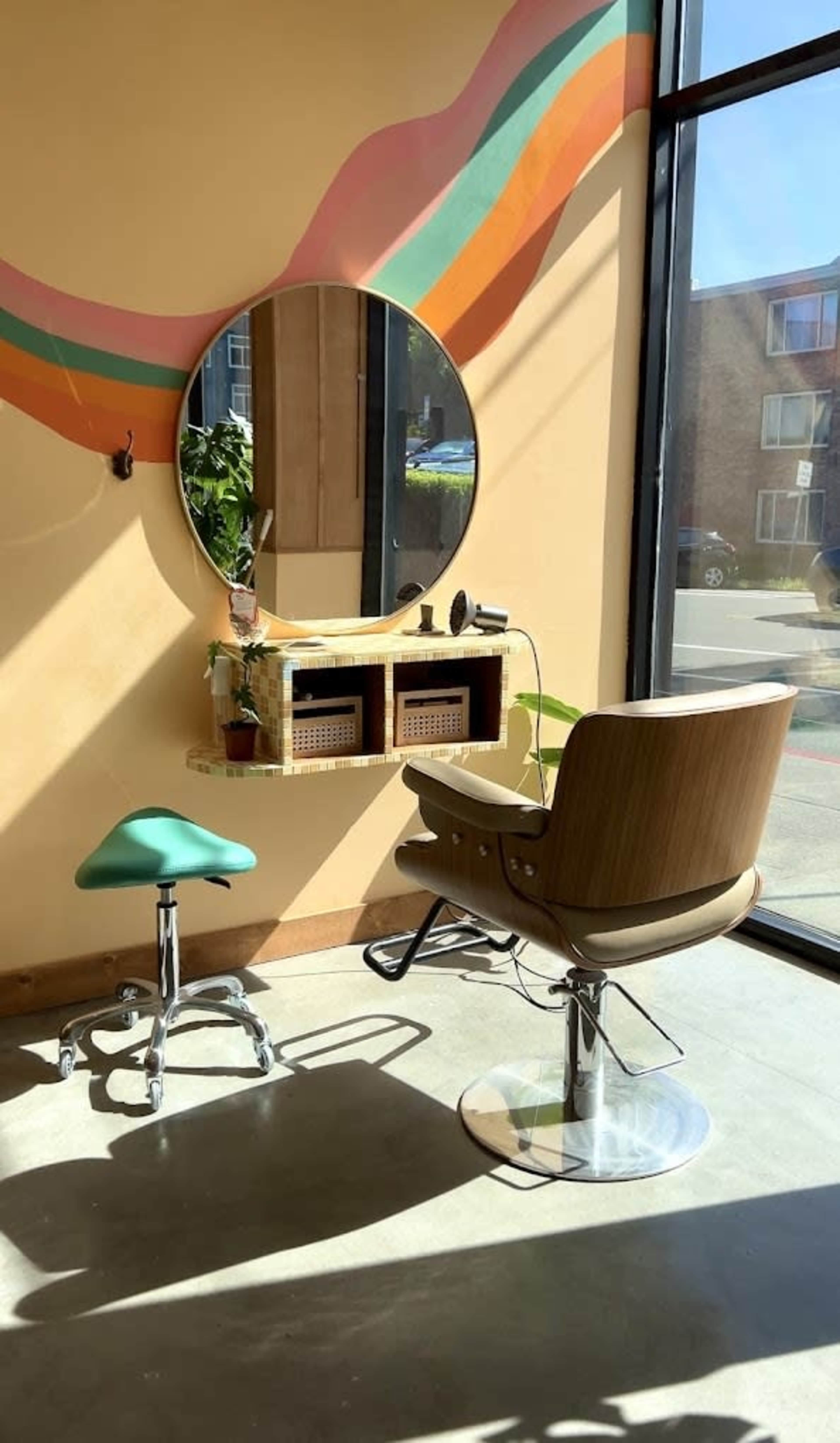 A salon interior features a large round mirror above a wooden shelf with storage baskets, accompanied by a styling chair and a stool.