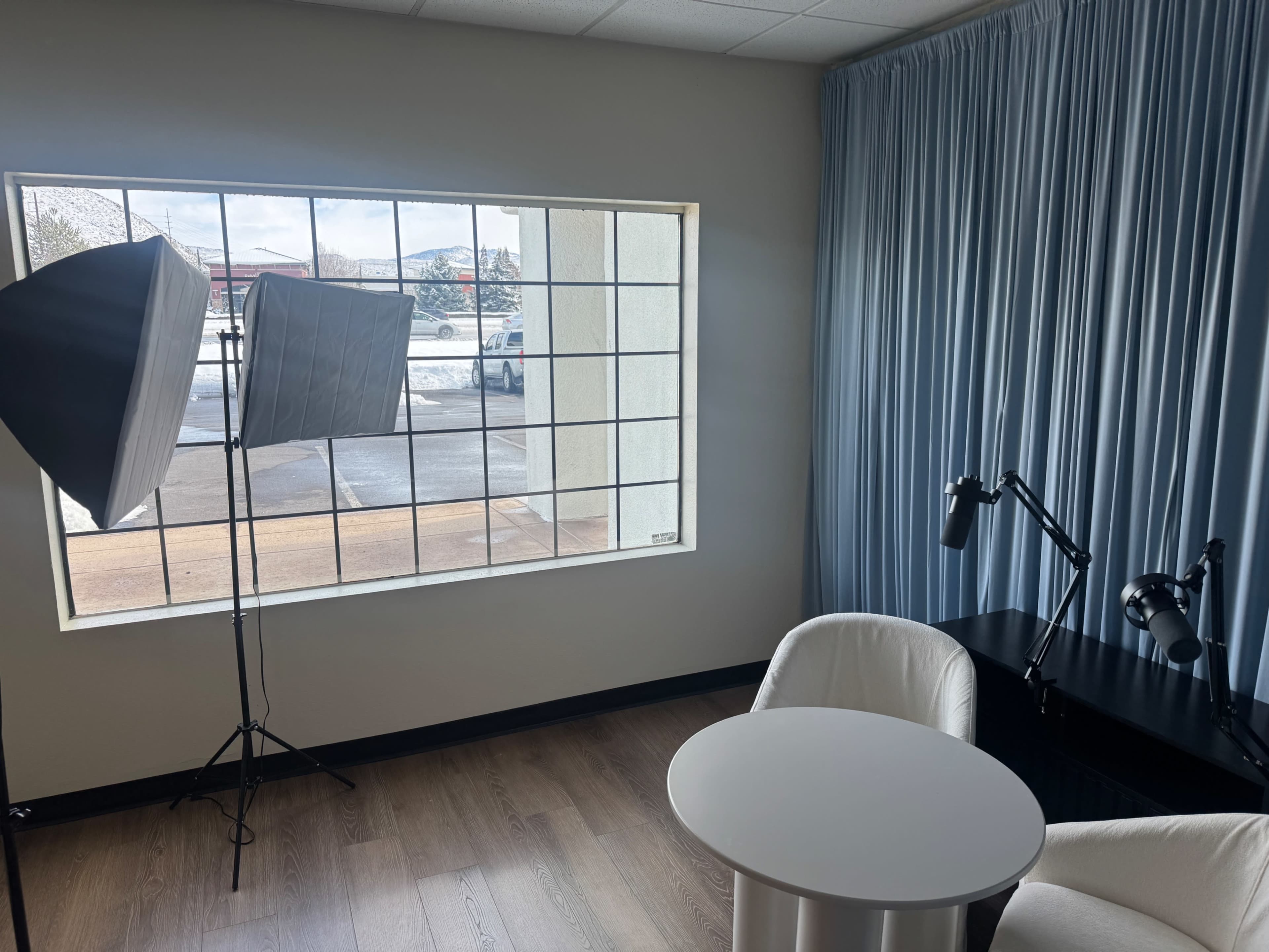 The image shows a well-lit room with a large window, two softbox lights, a small round table, and two white chairs.