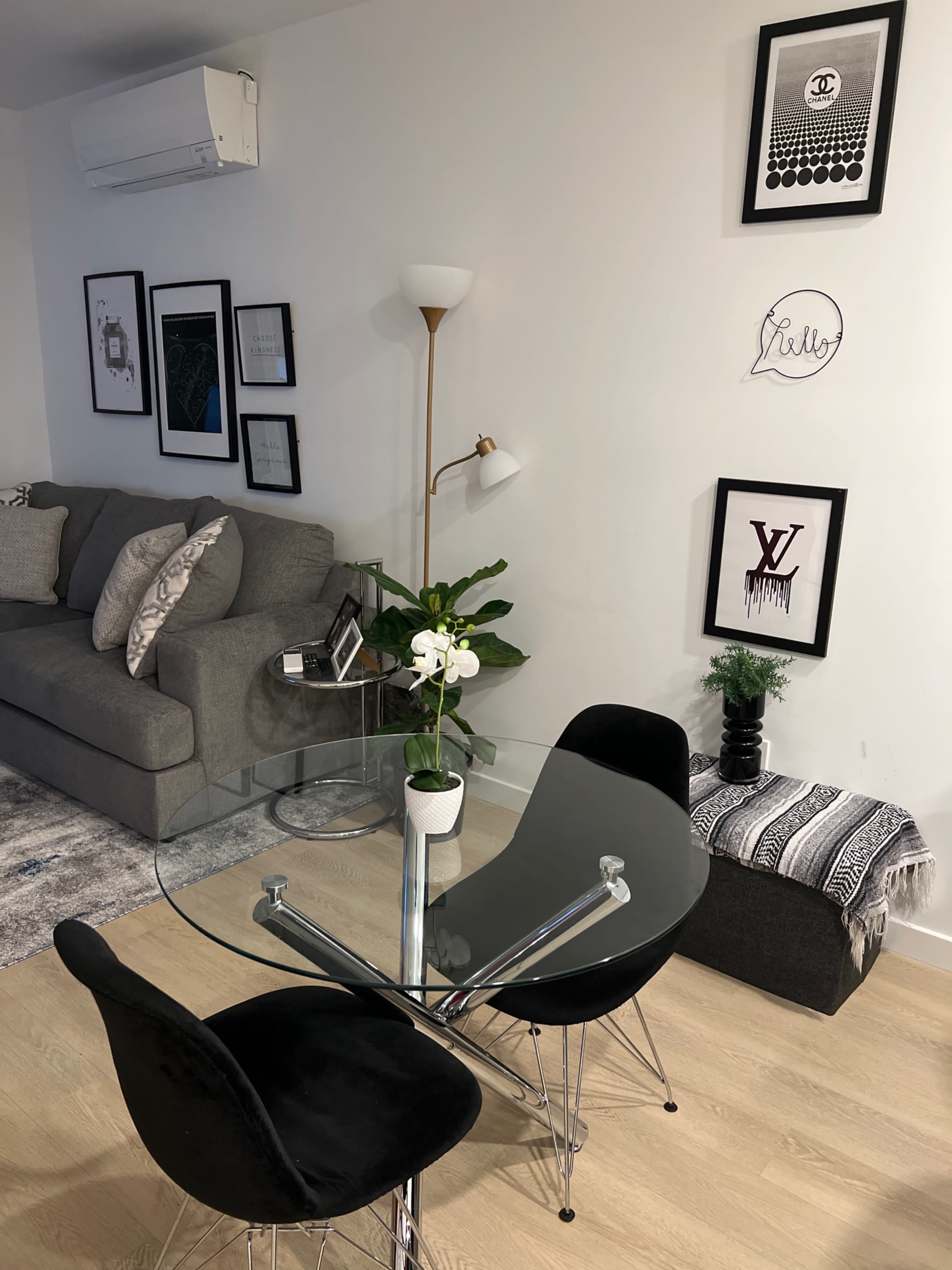 A modern dining area features a round glass table surrounded by two black chairs, with a gray sofa and various framed artworks on the walls.