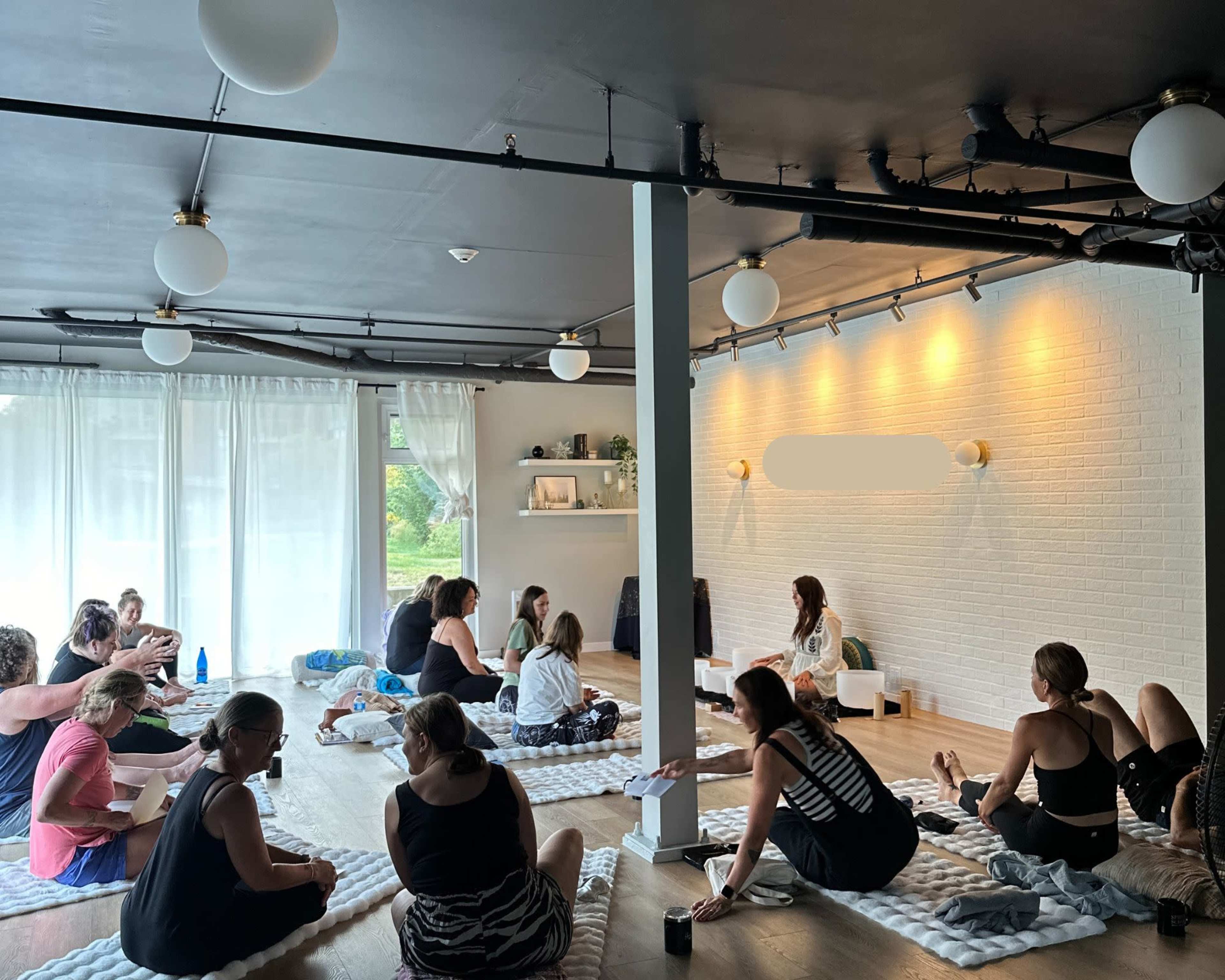 A group of people is seated on floor mats in a brightly lit room, with one person at the front speaking to the audience.