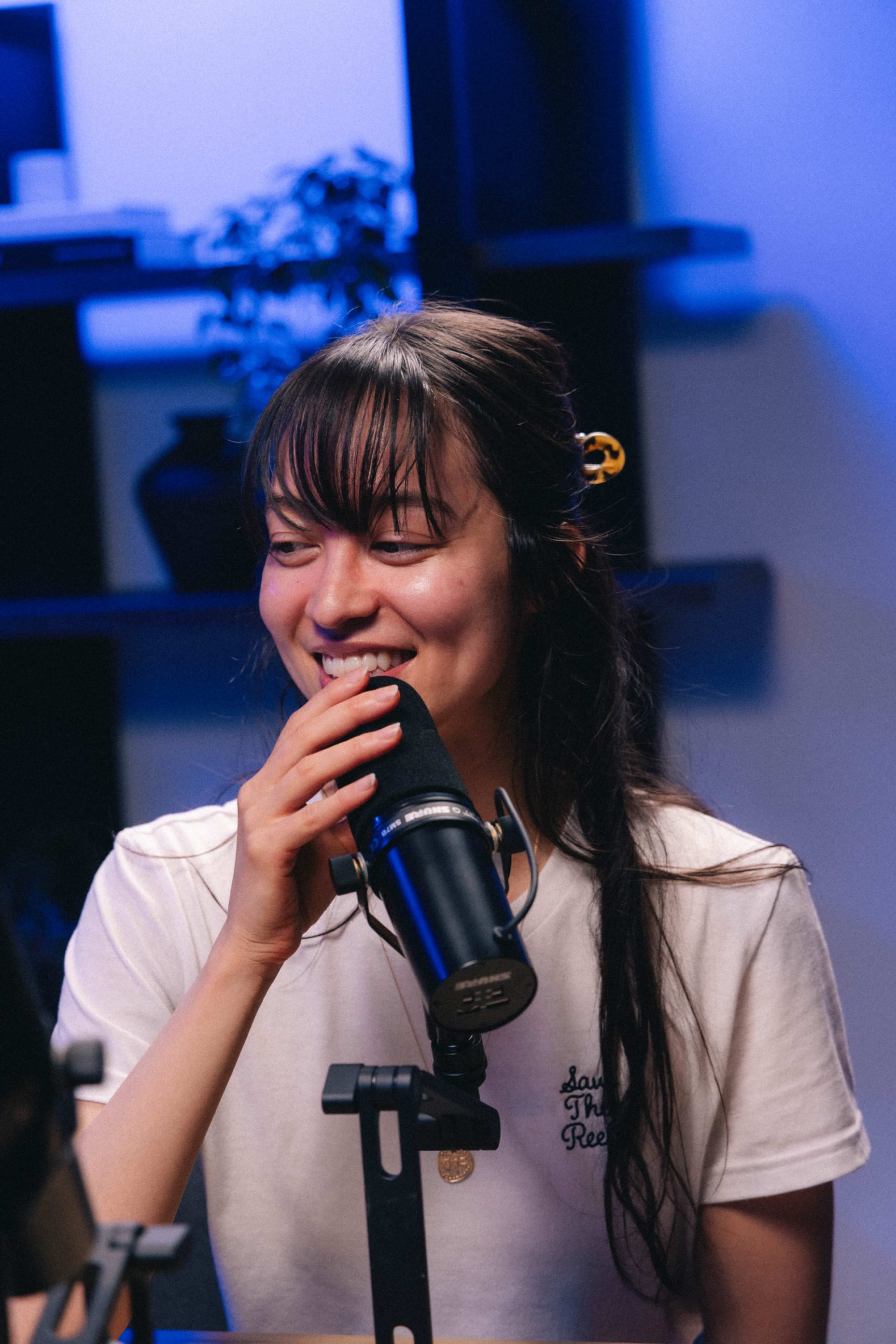 A young woman is speaking into a microphone while sitting at a table in a recording studio with blue lighting.