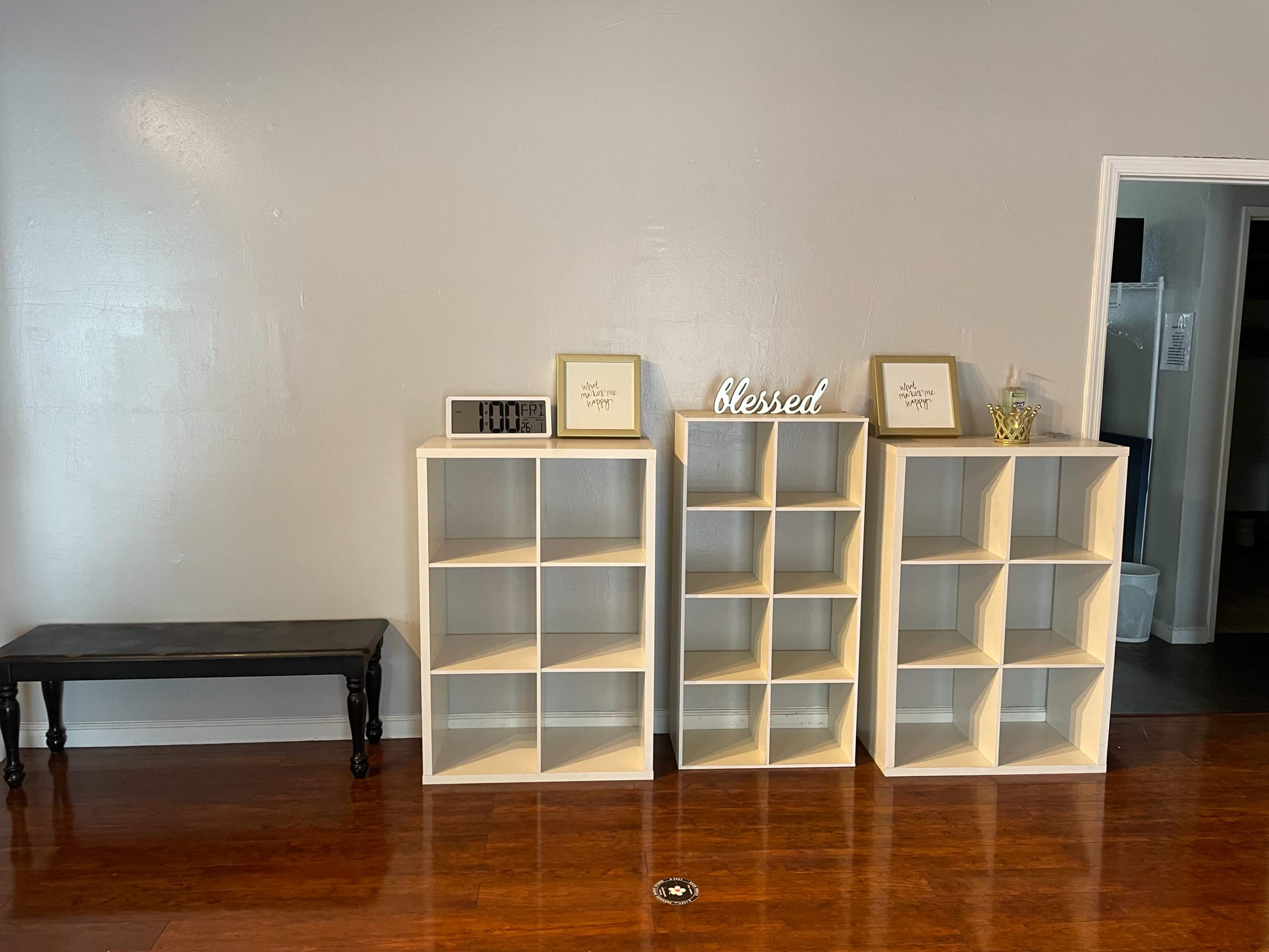 The image shows three wooden shelving units with decorative items on top, positioned against a gray wall, beside a black bench.