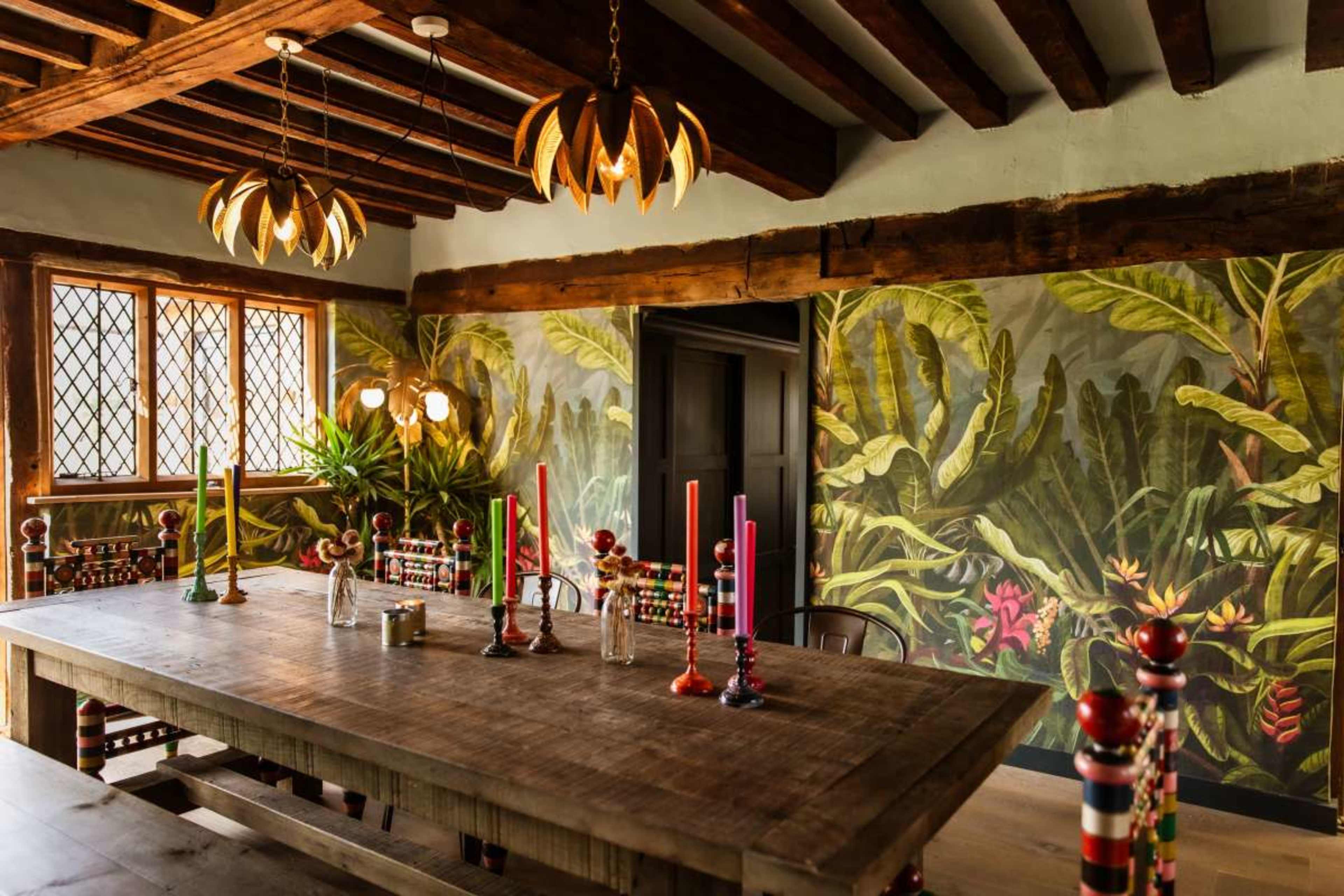 The image shows a dining room with a large wooden table, colorful candle holders, and a tropical mural on the wall.