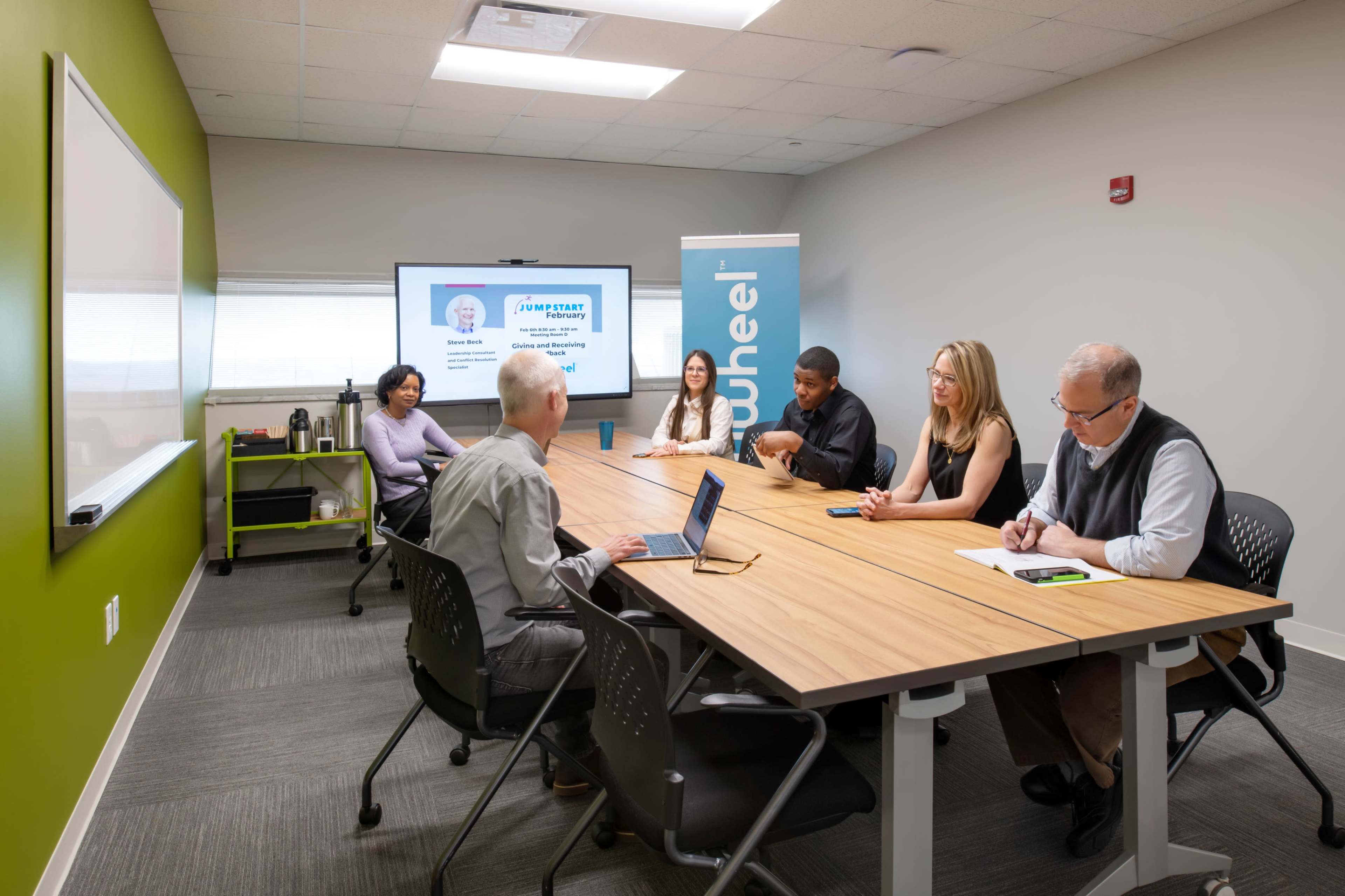 A group of six individuals is seated at a long conference table in a meeting room, with a presentation screen displaying a profile and the logo of a company named "wuneel."