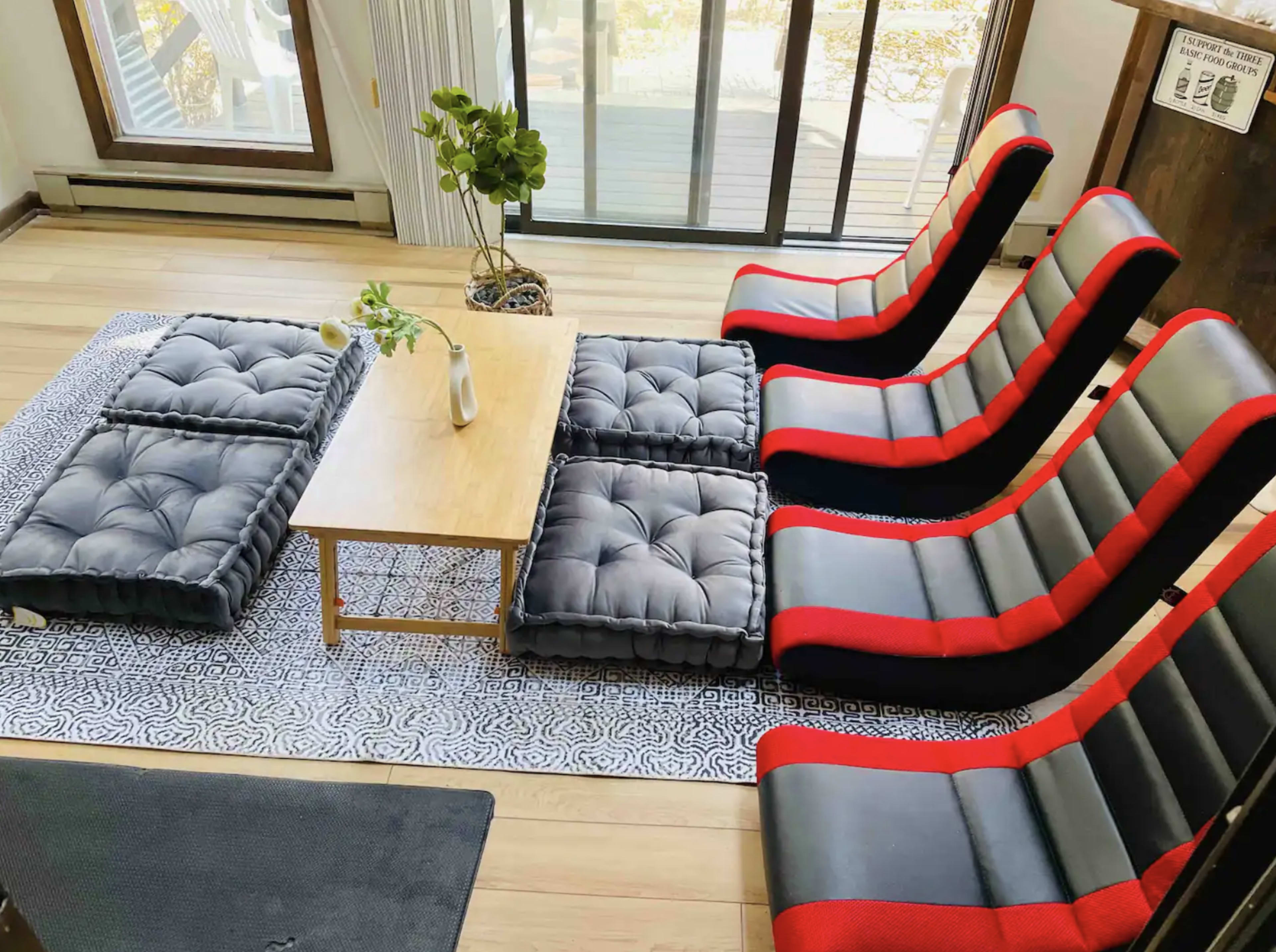 A modern living room with six lounge chairs arranged around a low wooden table, accompanied by floor cushions on a patterned rug.