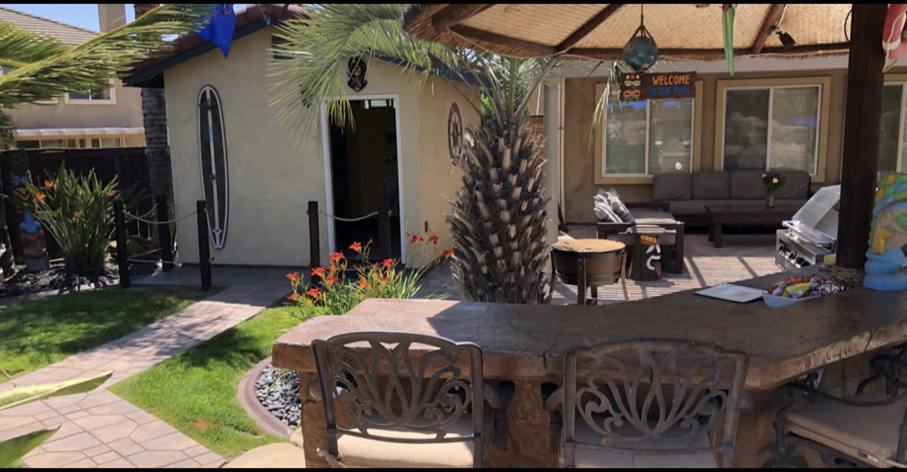 A backyard patio area with a stone bar, seating, palm trees, and a door leading into the house.