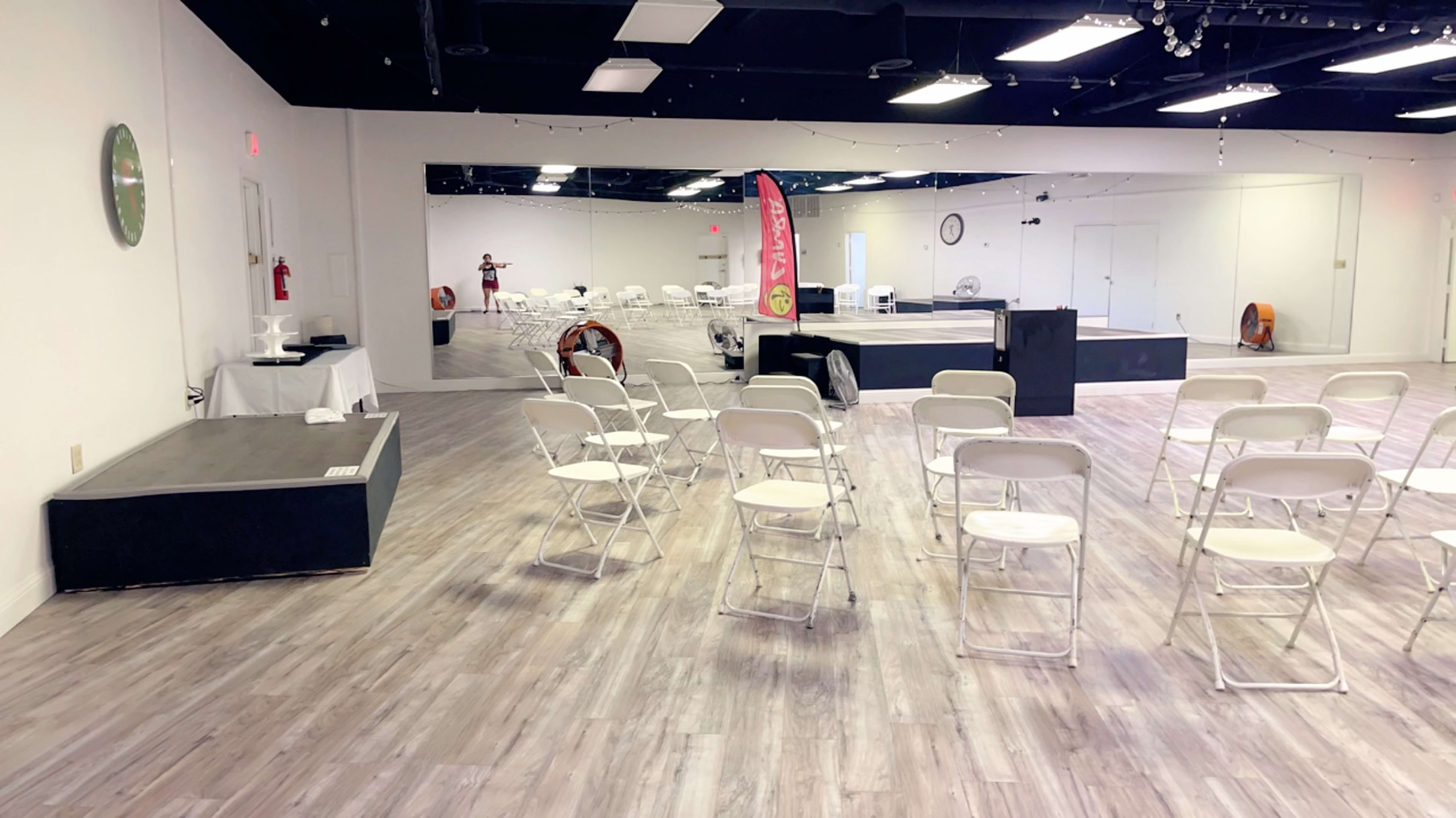 A spacious, empty indoor venue with rows of white folding chairs arranged in front of a stage and a mirrored wall.