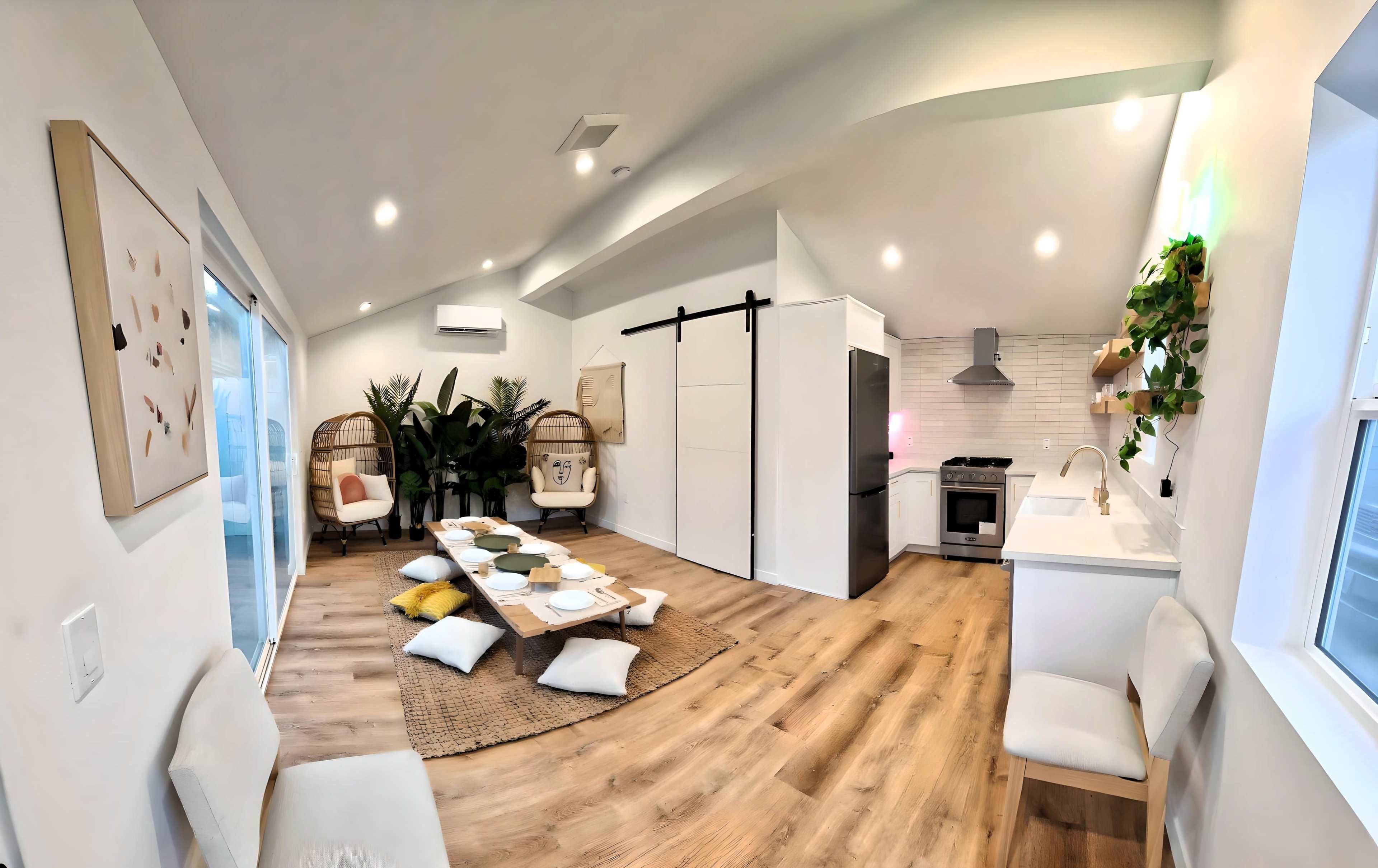 The image shows a bright, modern living space featuring a kitchen, dining area with floor cushions, and two woven chairs surrounded by indoor plants.