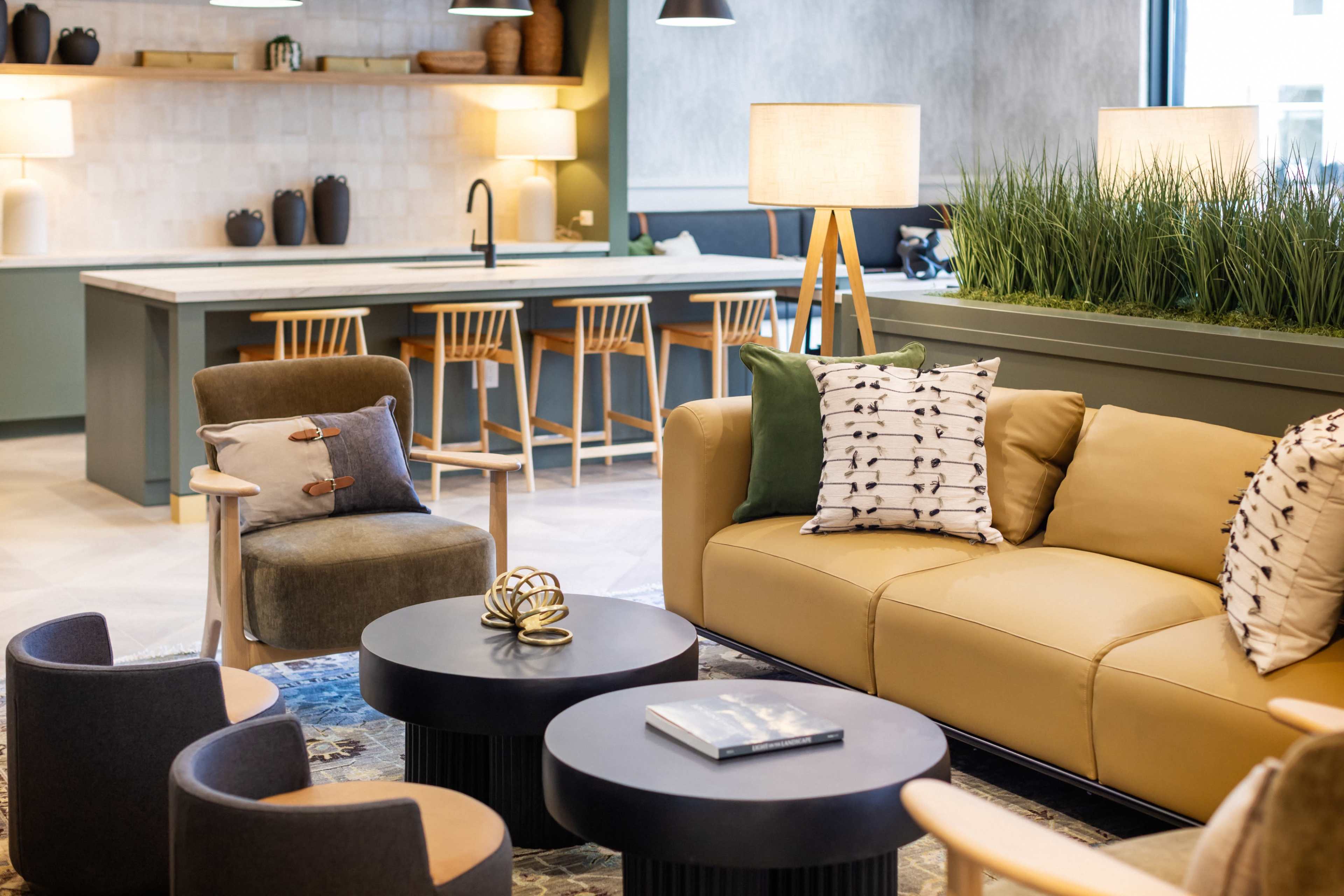 A modern living area features a beige sofa, dark coffee tables, wooden chairs, and a kitchen area in the background.